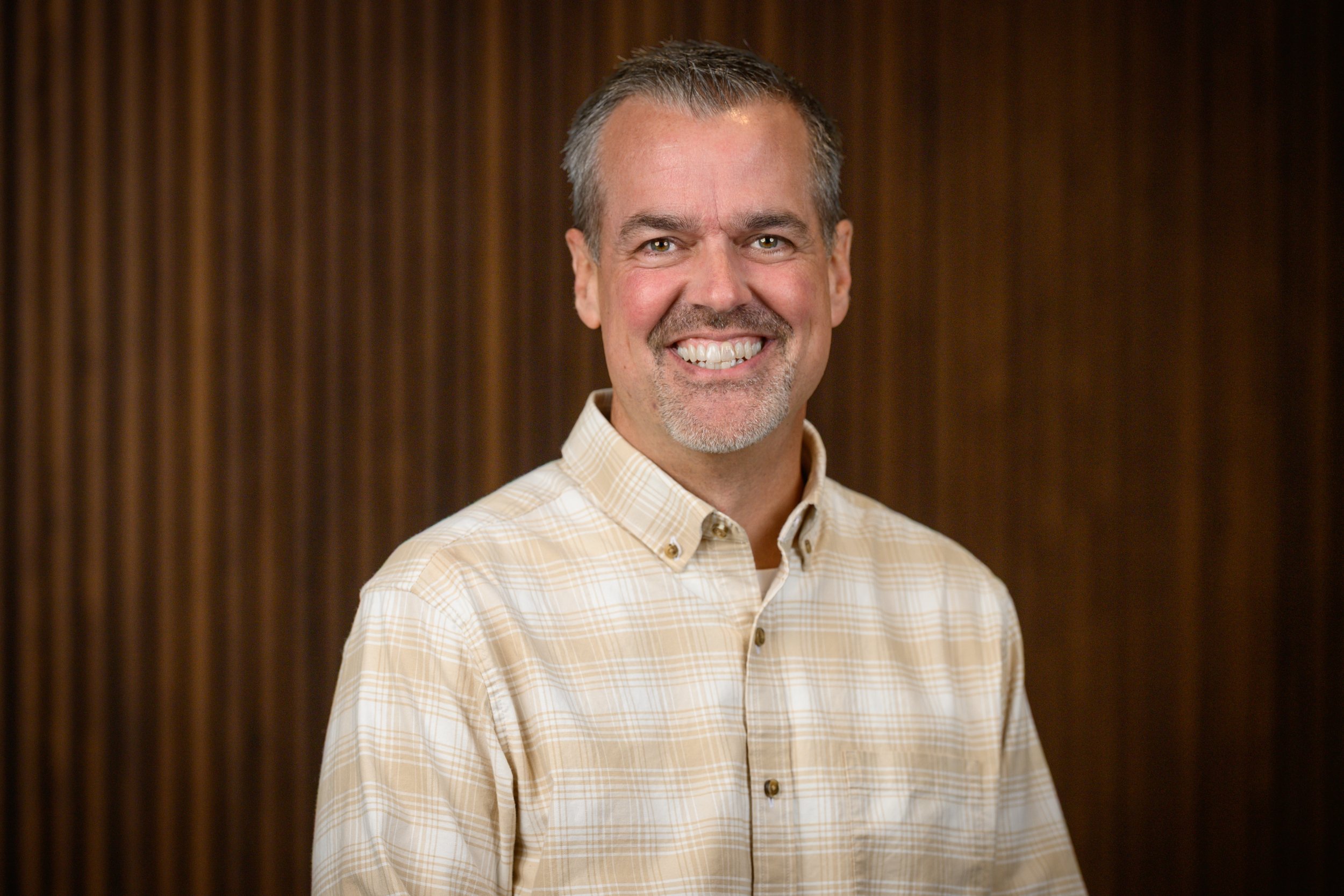 A smiling man with short gray hair and a beard wearing a light-colored plaid button-up shirt, standing in front of a wooden background.
