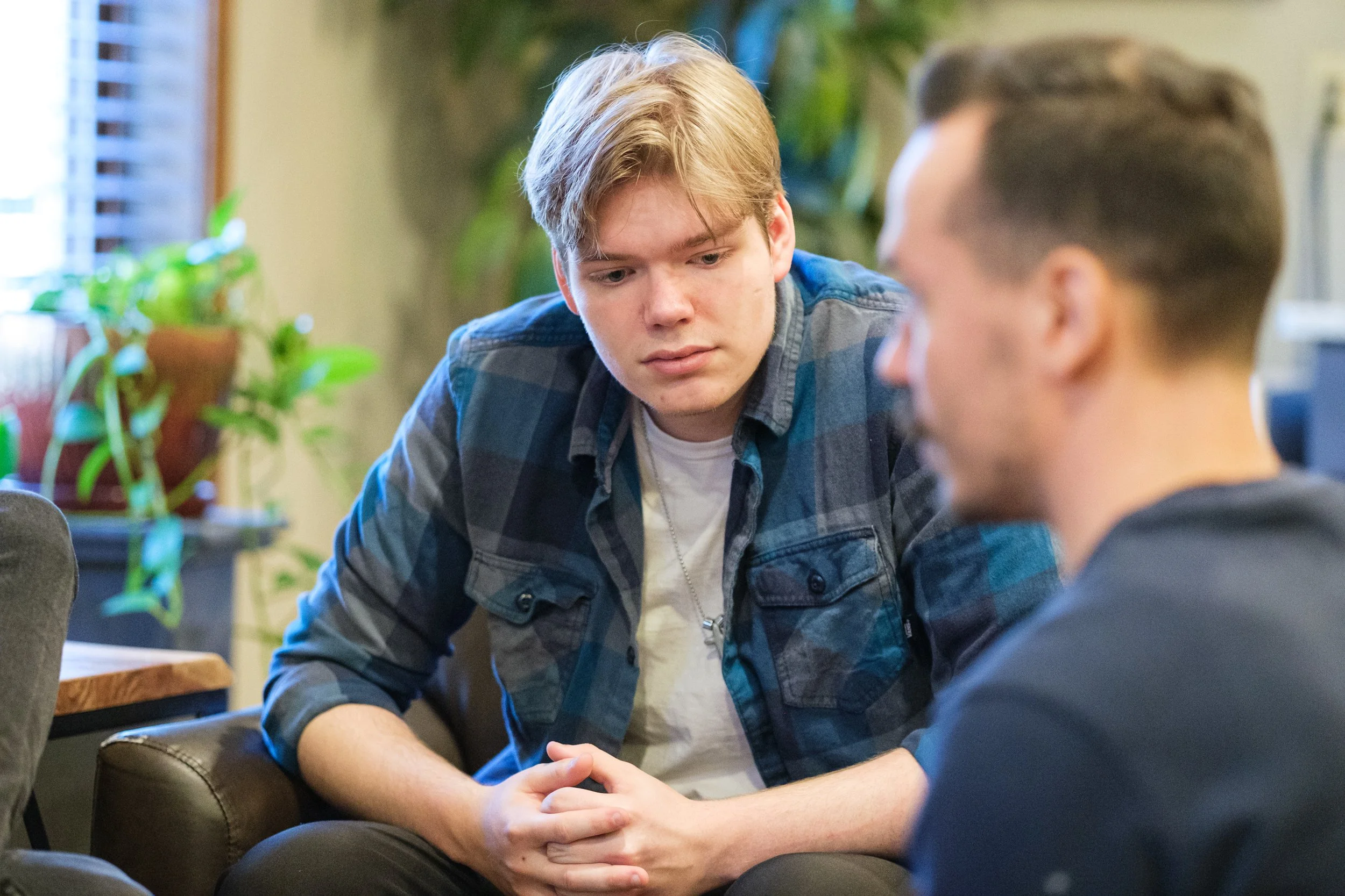 Two young men sitting close together in a cozy indoor setting, engaged in a serious conversation. One has light brown hair, wearing a blue flannel shirt over a white T-shirt, while the other, with darker hair, is partially visible.