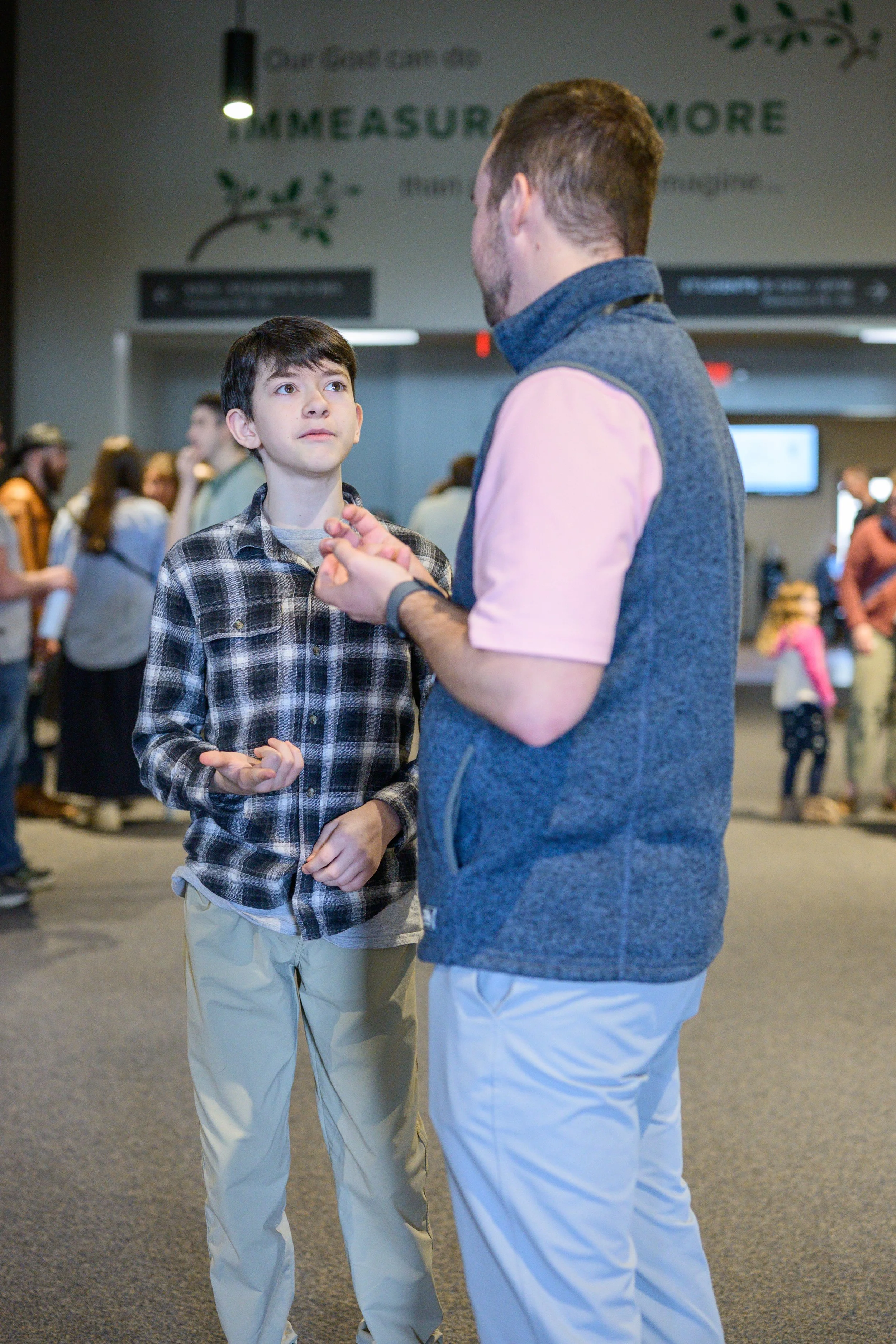 A young boy with dark hair, wearing a plaid shirt and light-colored pants, looks at an adult man with light brown hair, wearing a sleeveless vest over a pink shirt and white pants, in a busy indoor setting.