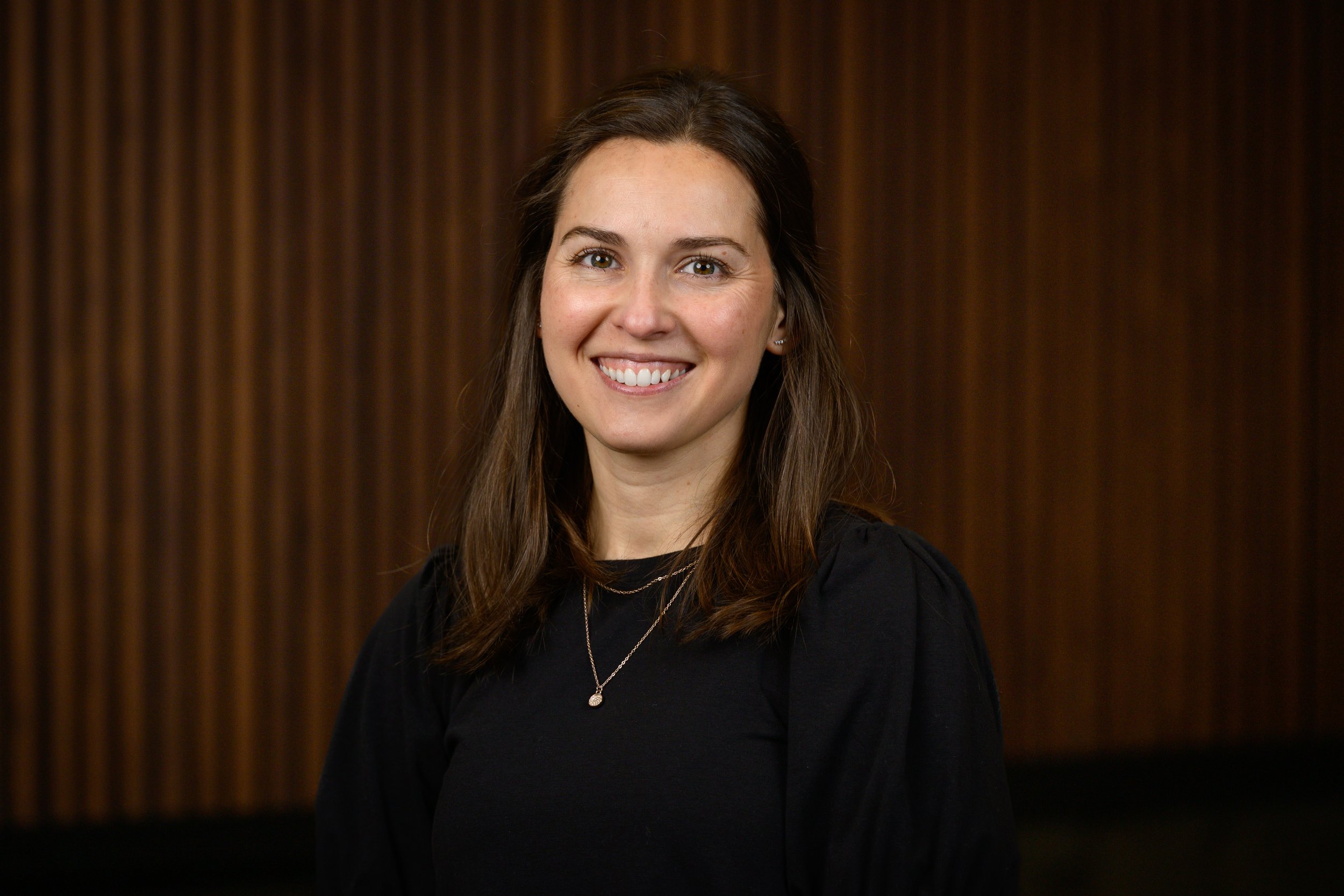Portrait of a young woman with long brown hair smiling, wearing a black top, standing against a dark wooden background.