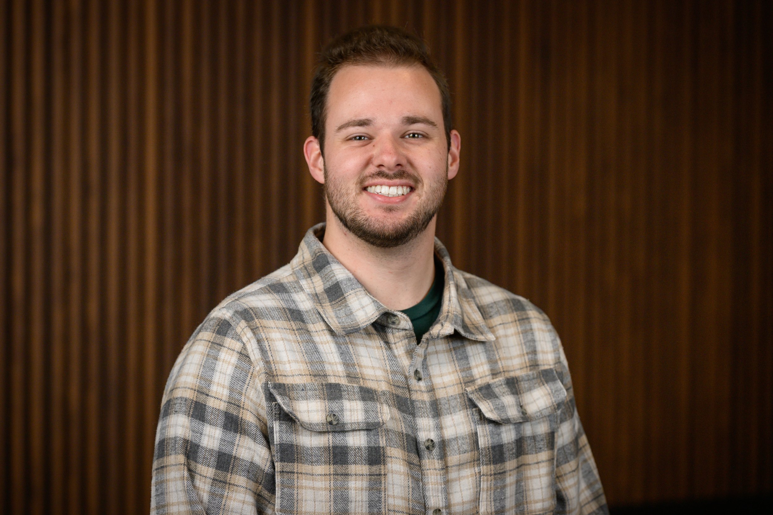 A smiling young man with short brown hair, facial hair, and a plaid shirt standing in front of a wooden background.