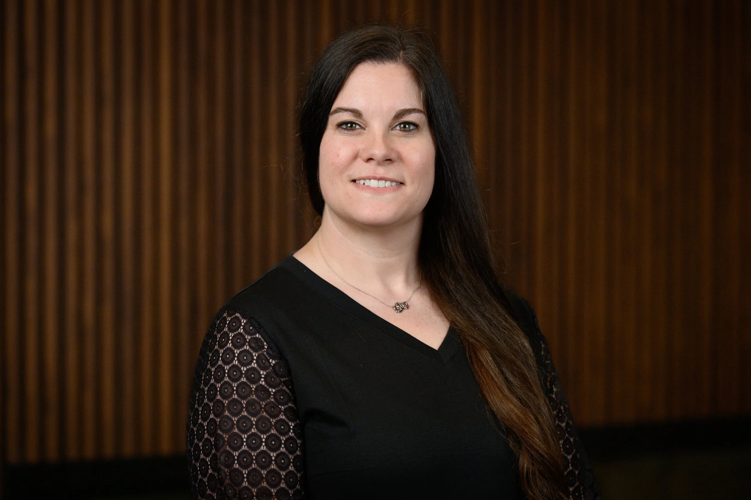 A woman with long, dark hair and light skin wearing a black top with lace sleeves and a silver necklace, standing in front of a wooden panel background.