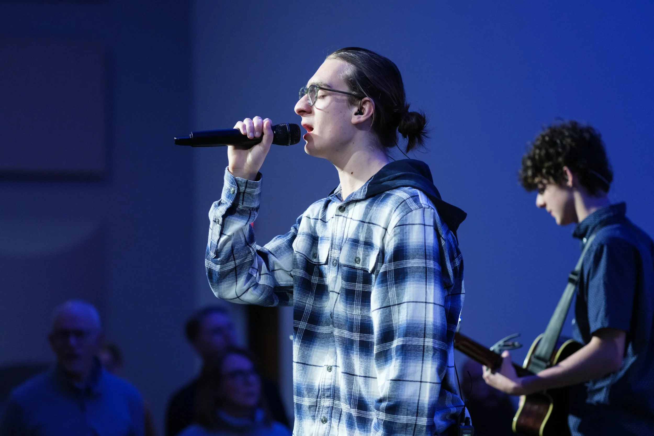 A young man with glasses singing into a microphone and wearing a plaid shirt on stage. Another person beside him is playing guitar. The background is blue and dark, suggesting an indoor performance or rehearsal.