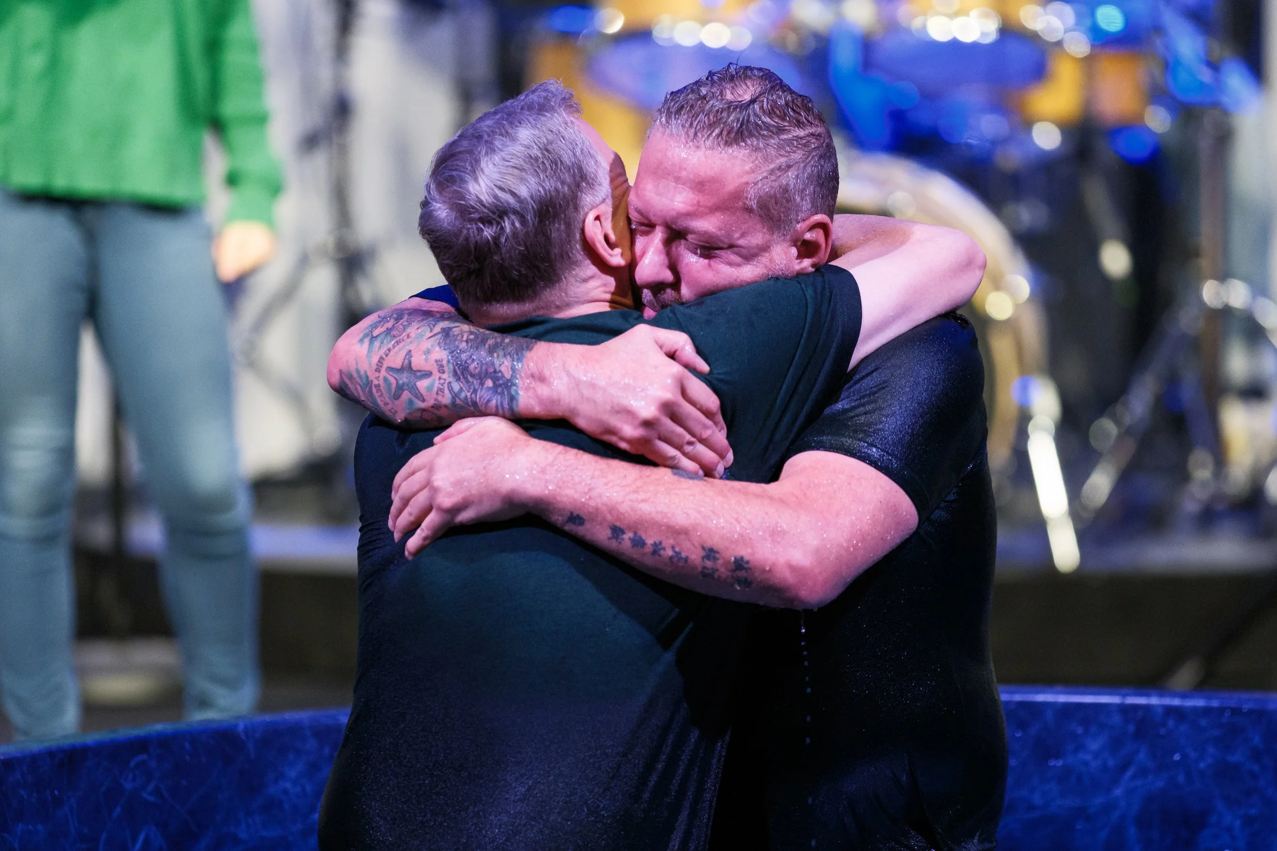 Two men hugging tightly after a baptism ceremony, with water on their skin and joyful expressions.