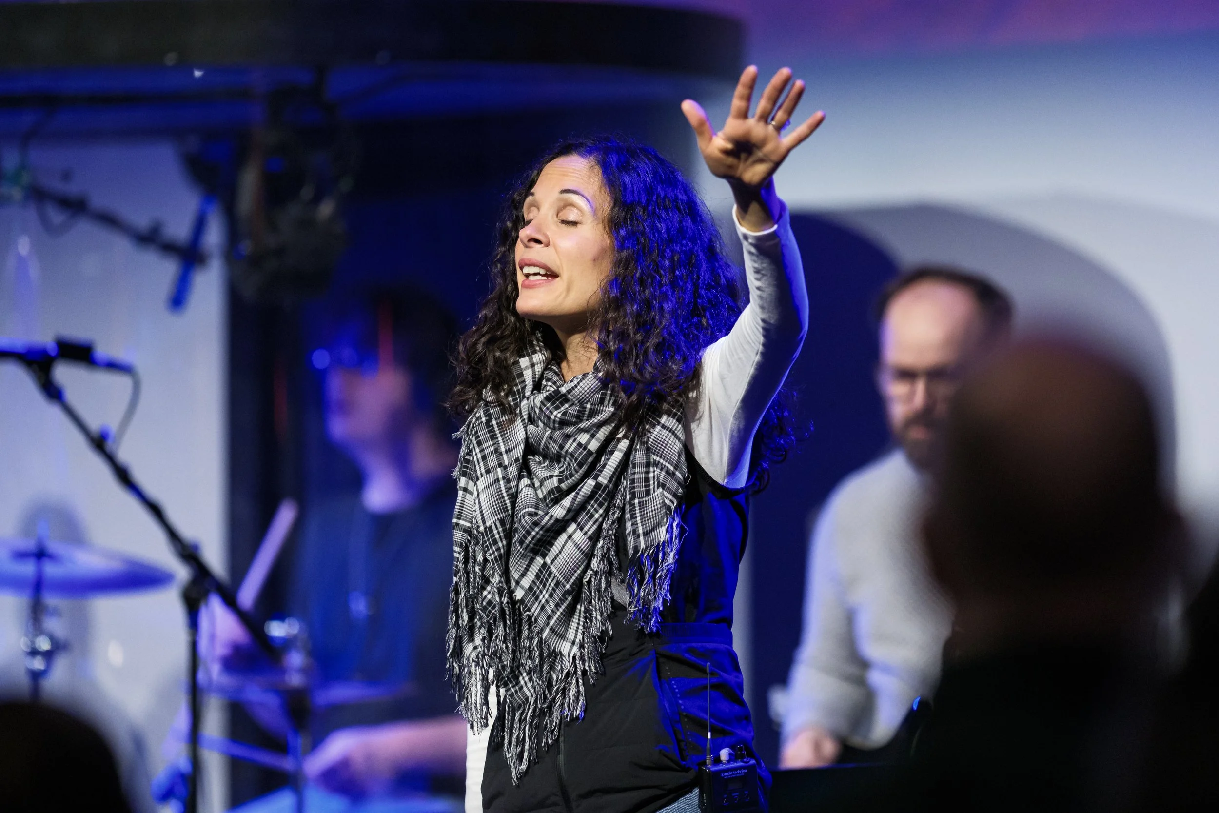 A woman with long curly hair singing or speaking with her eyes closed and her hand raised in the air, in a dimly lit setting with purple and blue lighting.