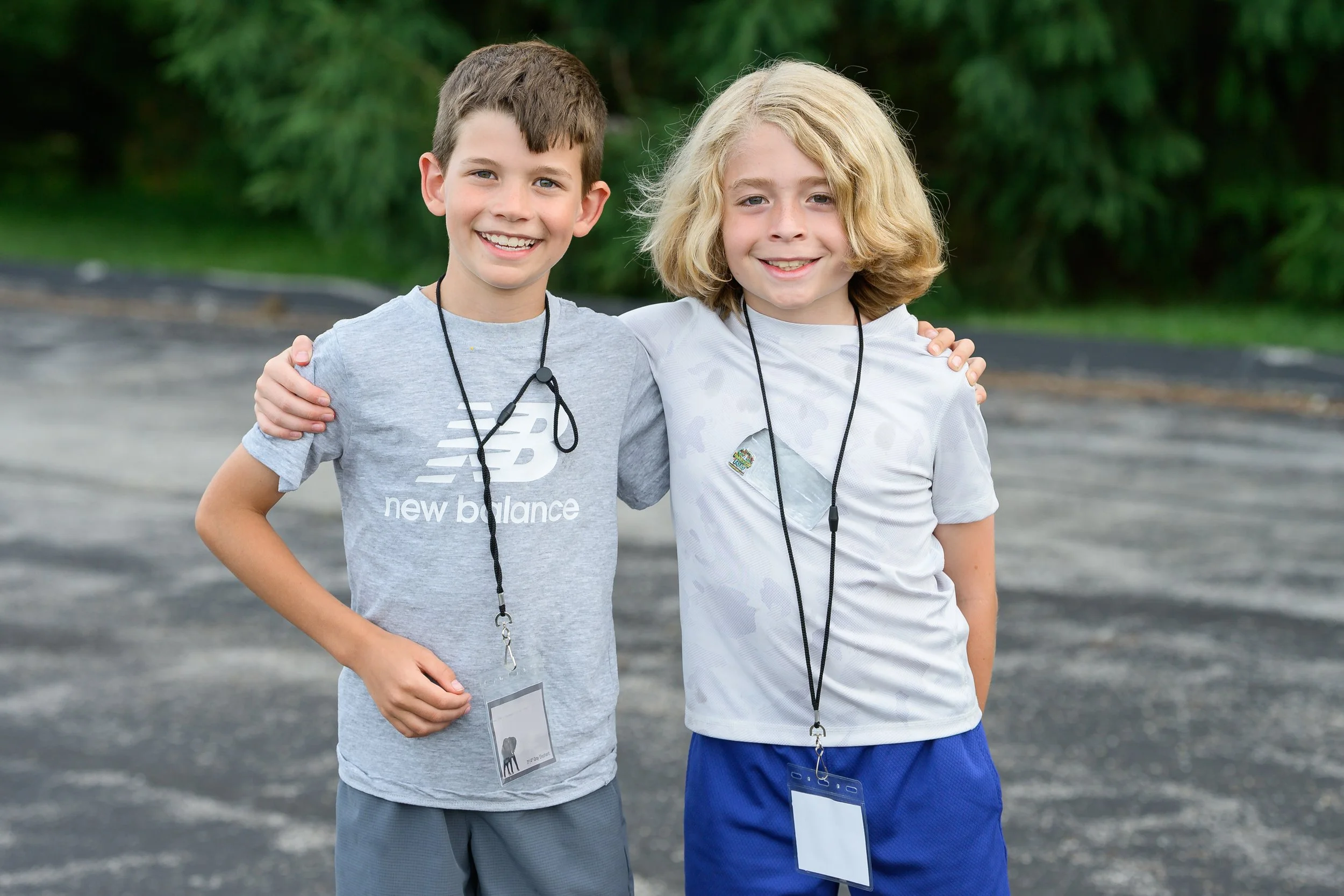 Two smiling boys with lanyards around their necks standing outdoors on a paved area with greenery in the background, embracing each other.
