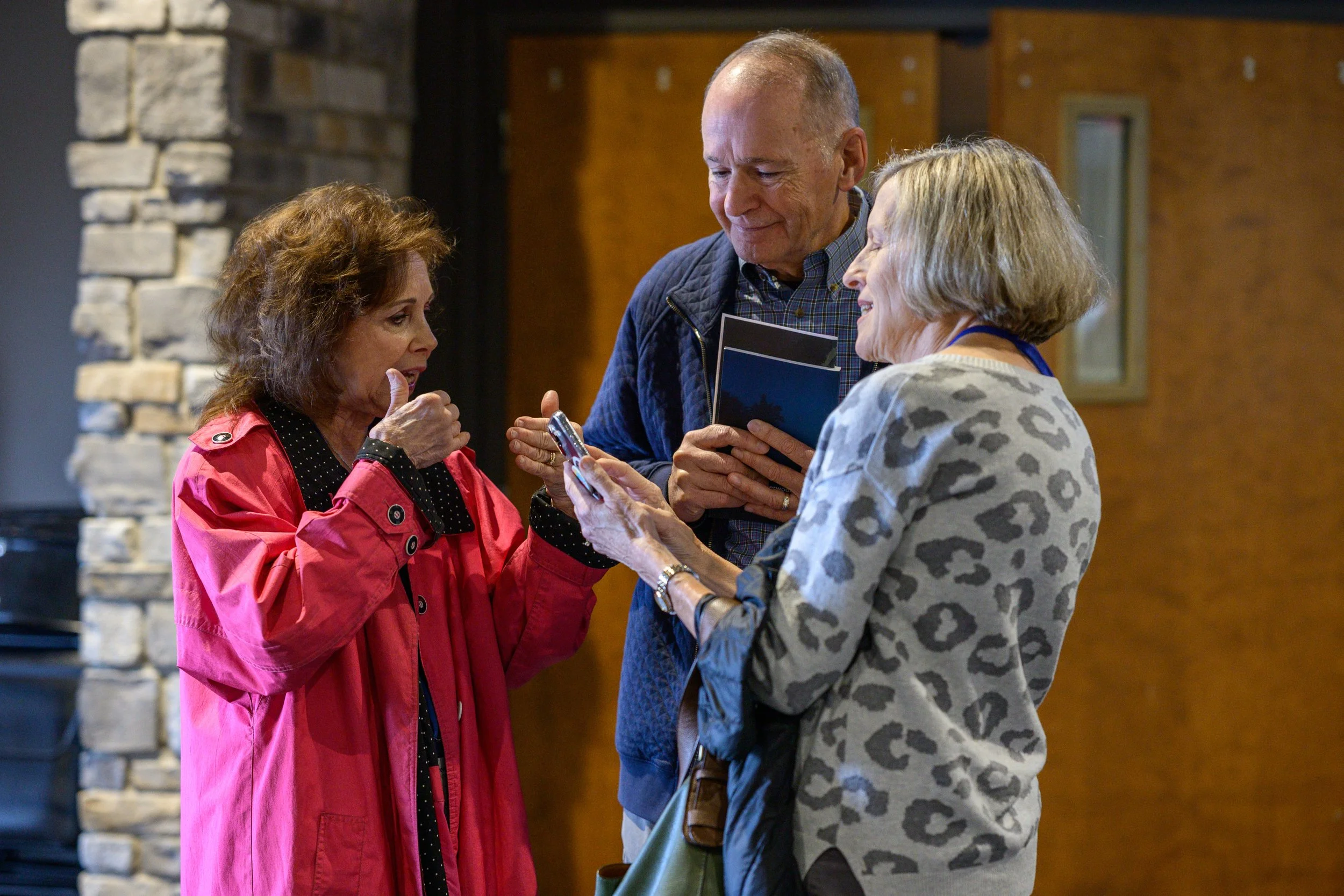 Three older adults standing close together and smiling, looking at a smartphone held by the woman on the right in a room with brick and wood walls.