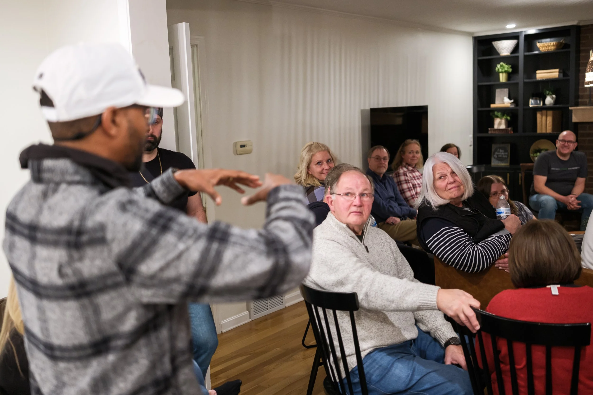 A group of people sitting and listening to a speaker in an indoor setting, with one man speaking at the front wearing a white cap and gray hoodie, and the audience showing attentive expressions.