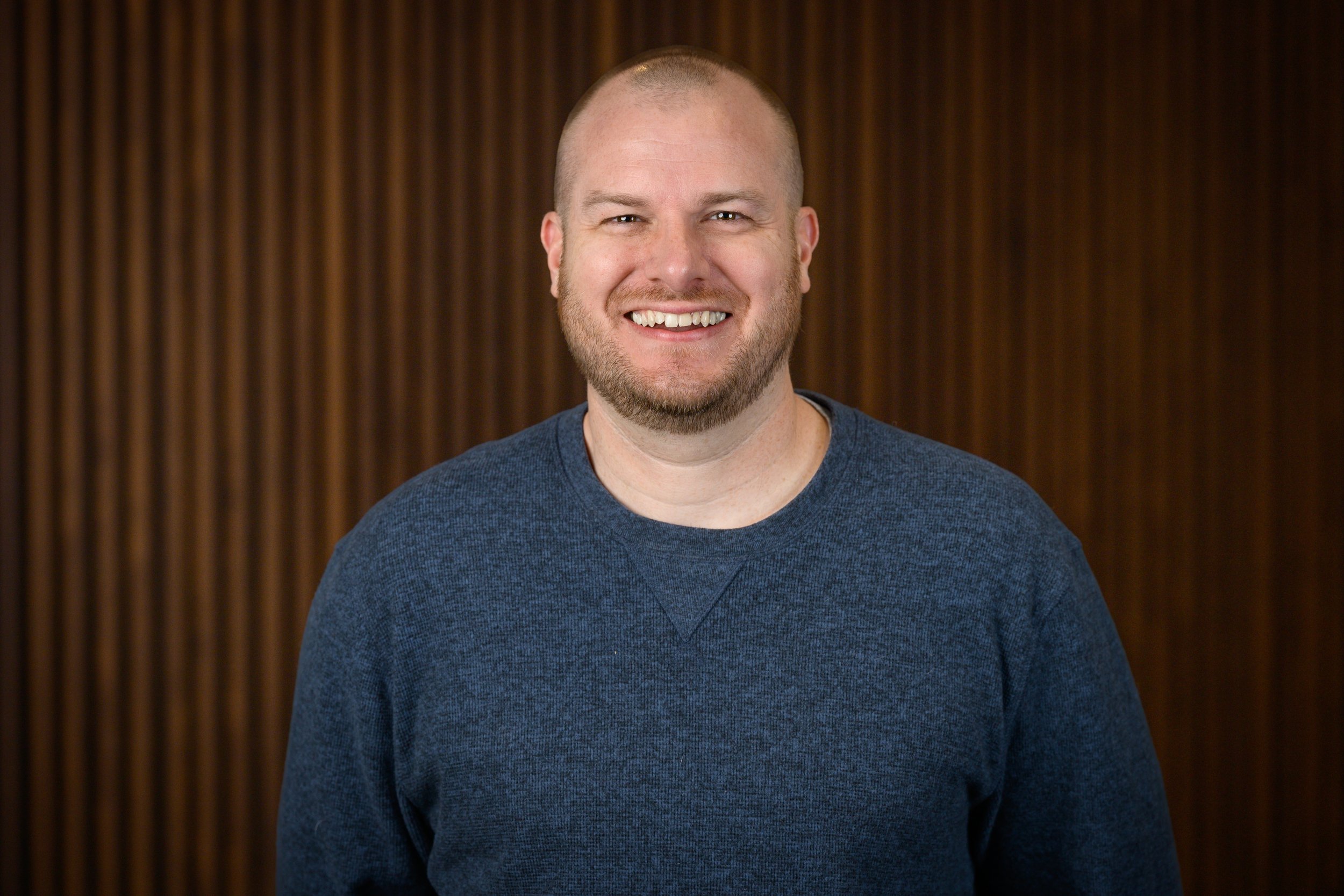 A smiling man with a bald head and a beard, wearing a dark gray sweater, standing in front of a wooden paneled background.