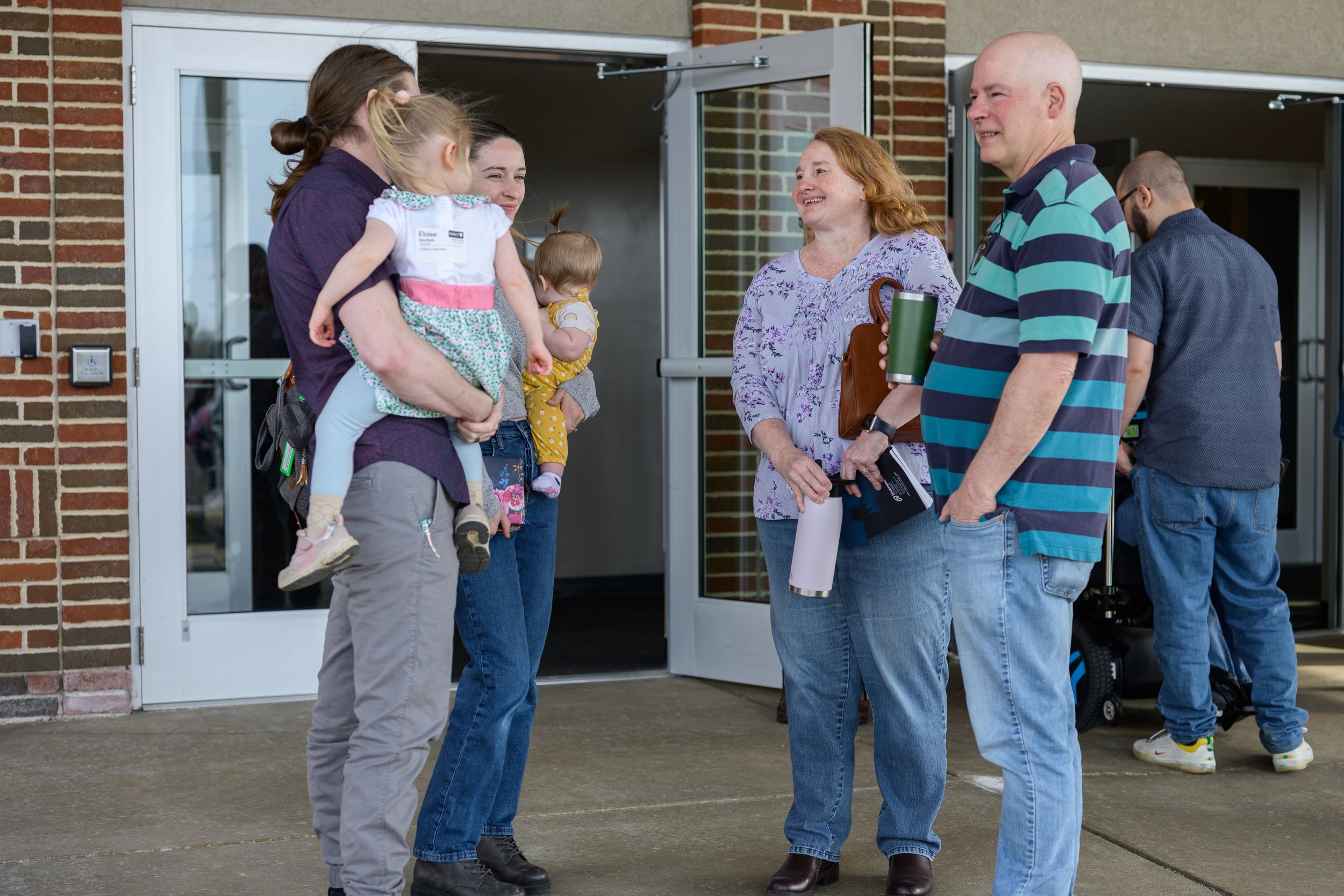 A group of people greeting each other outside a building, some holding children and drinks.