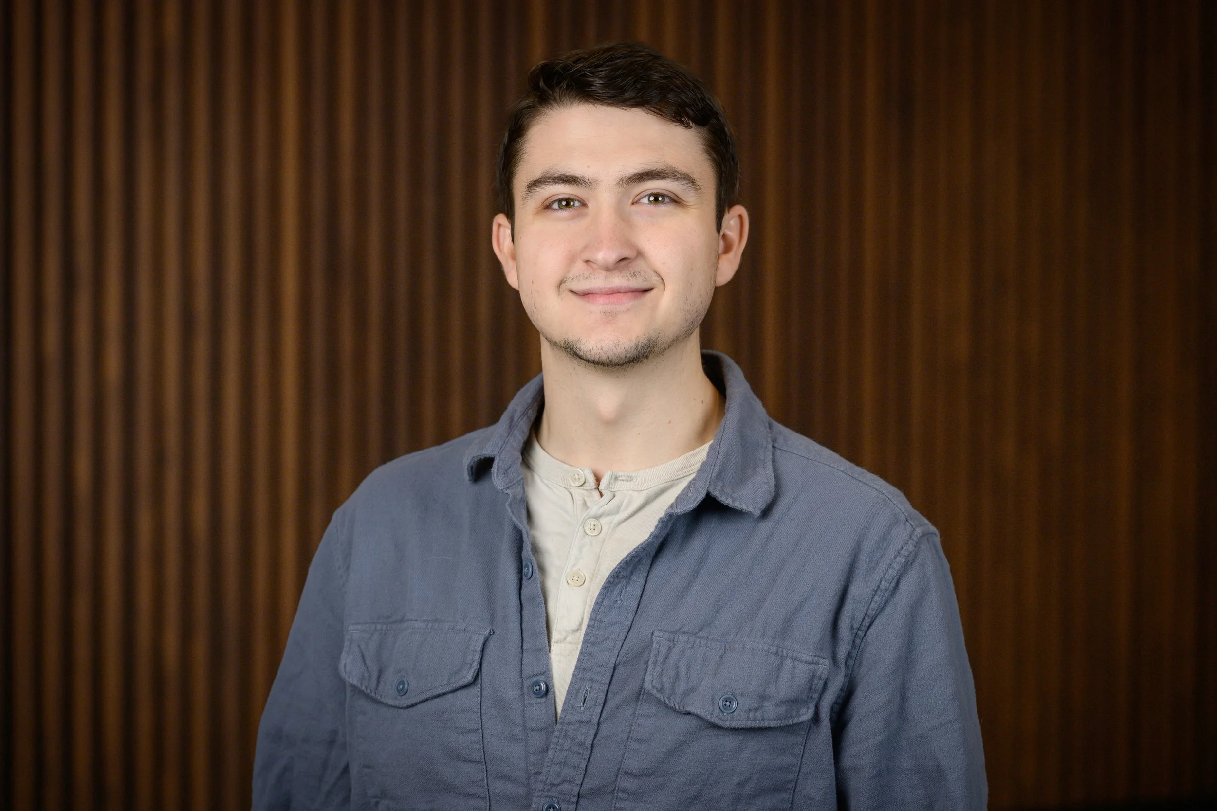 Portrait of a young man with short dark hair, light skin, wearing a blue button-up shirt over a light-colored undershirt, standing against a wooden-paneled background.