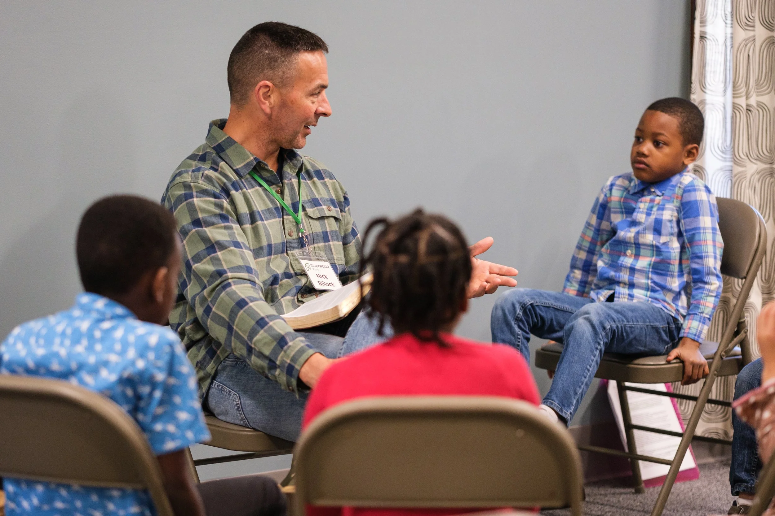 A man with short hair and a beard, wearing a plaid shirt and a name tag, is talking to a young boy with short hair, wearing a plaid shirt, sitting on a chair. Other children are seated around them, and the scene appears to be in a classroom or community center with a plain gray wall and curtain in the background.