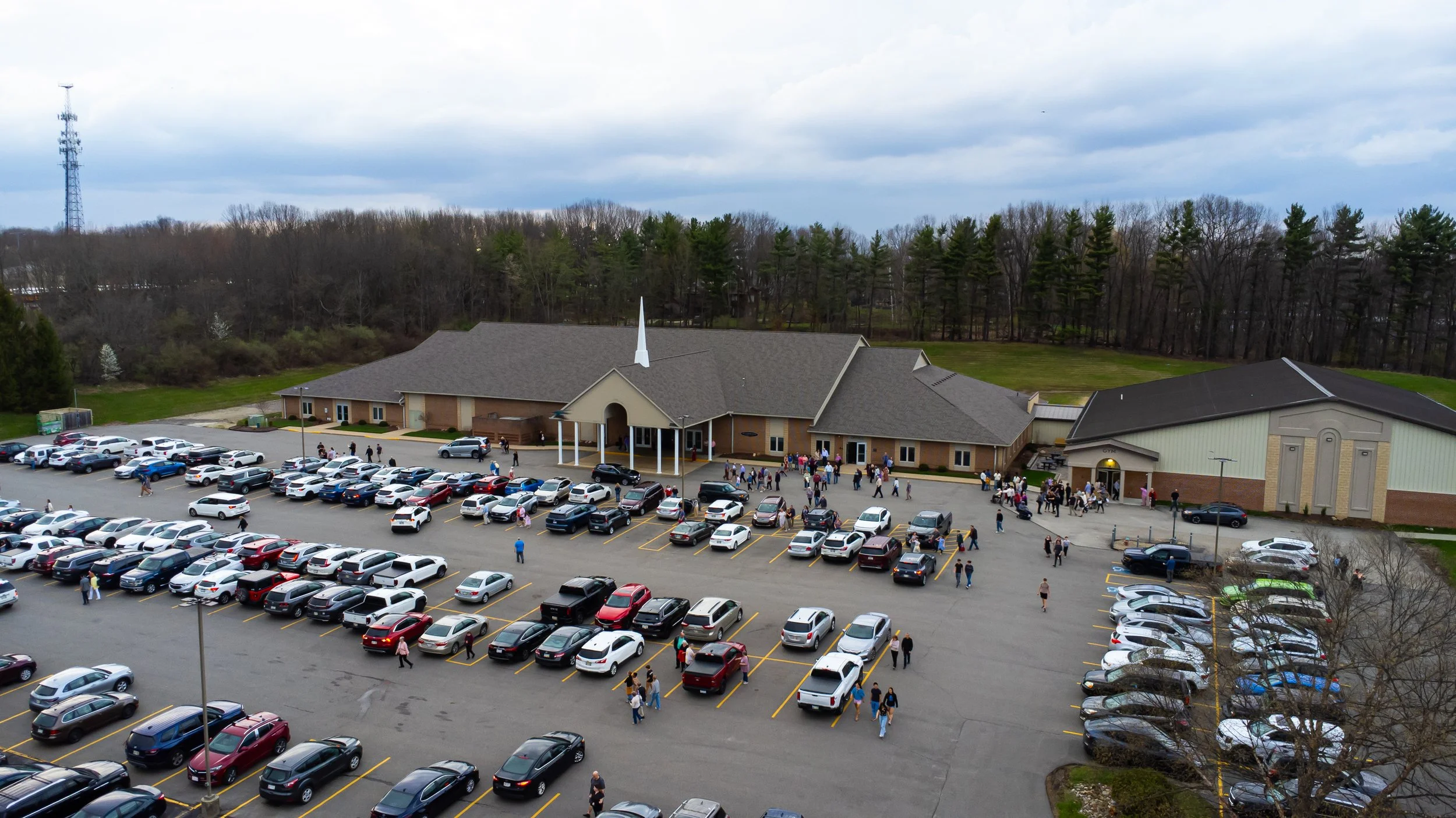 A large brick church with a tall white steeple, surrounded by a parking lot filled with cars and groups of people walking around, with a background of trees and cloudy sky.