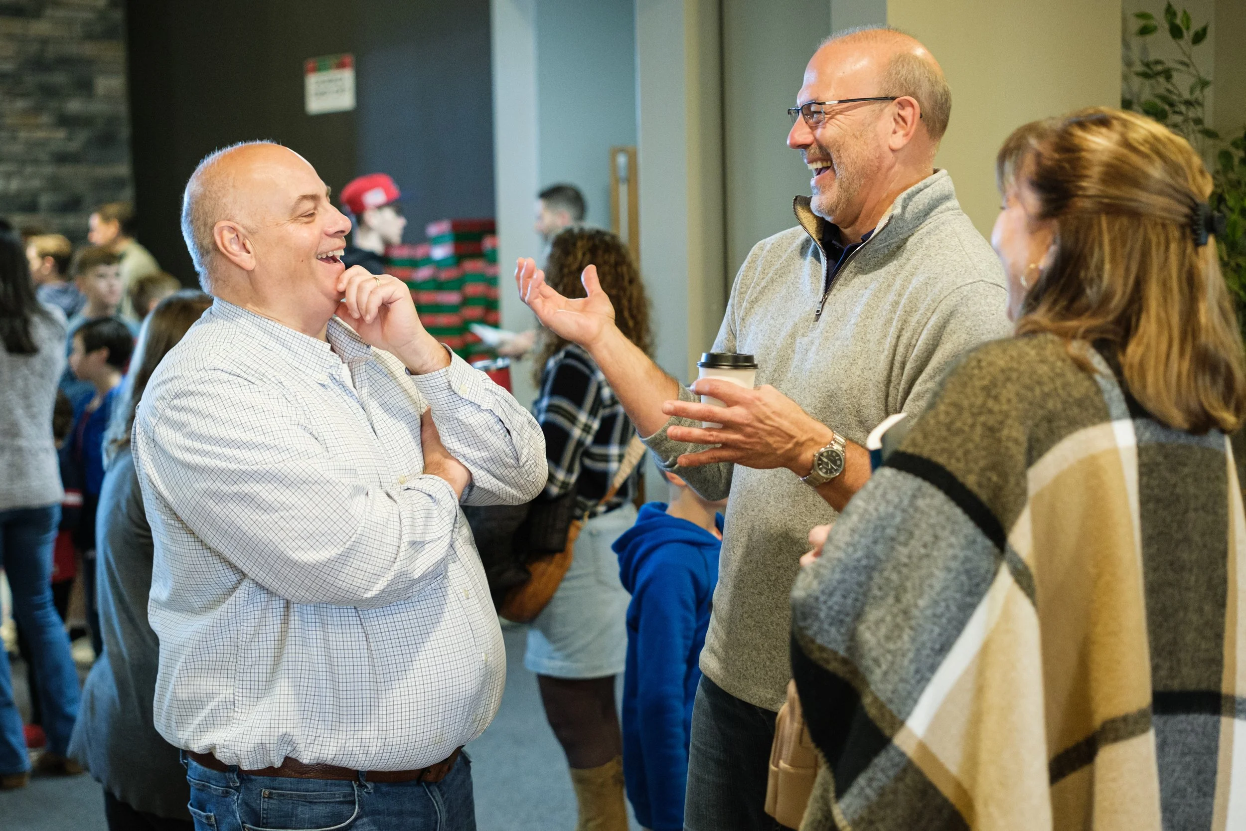 Two men are laughing and talking to each other at a joyful social gathering, with other people in the background.