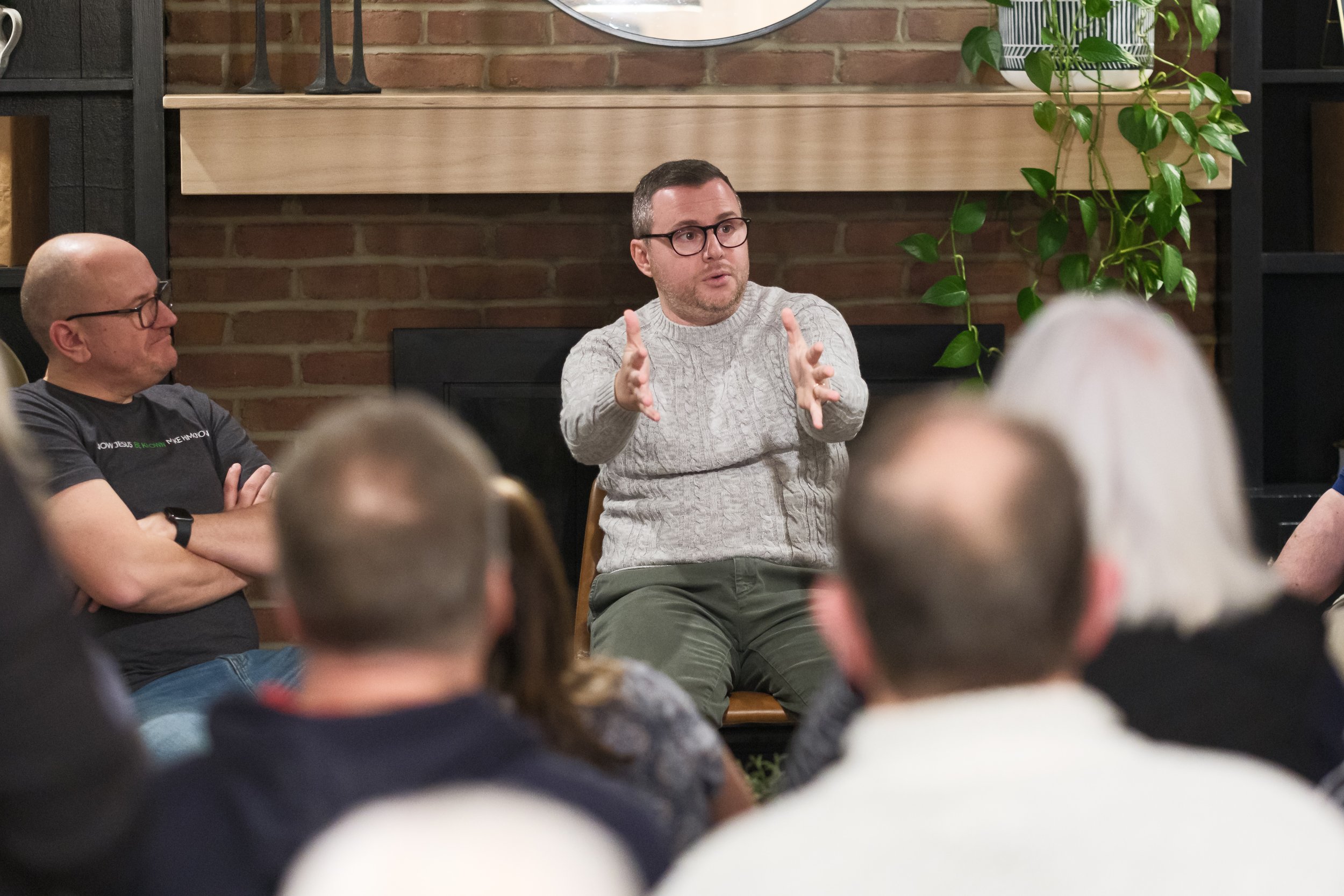 A man wearing glasses, a light gray sweater, and olive green pants is speaking and gesturing with his hands during a discussion. He is sitting in front of a brick wall with a large round mirror, some greenery, and decorative items on shelves. Several other people are seated around him, listening.