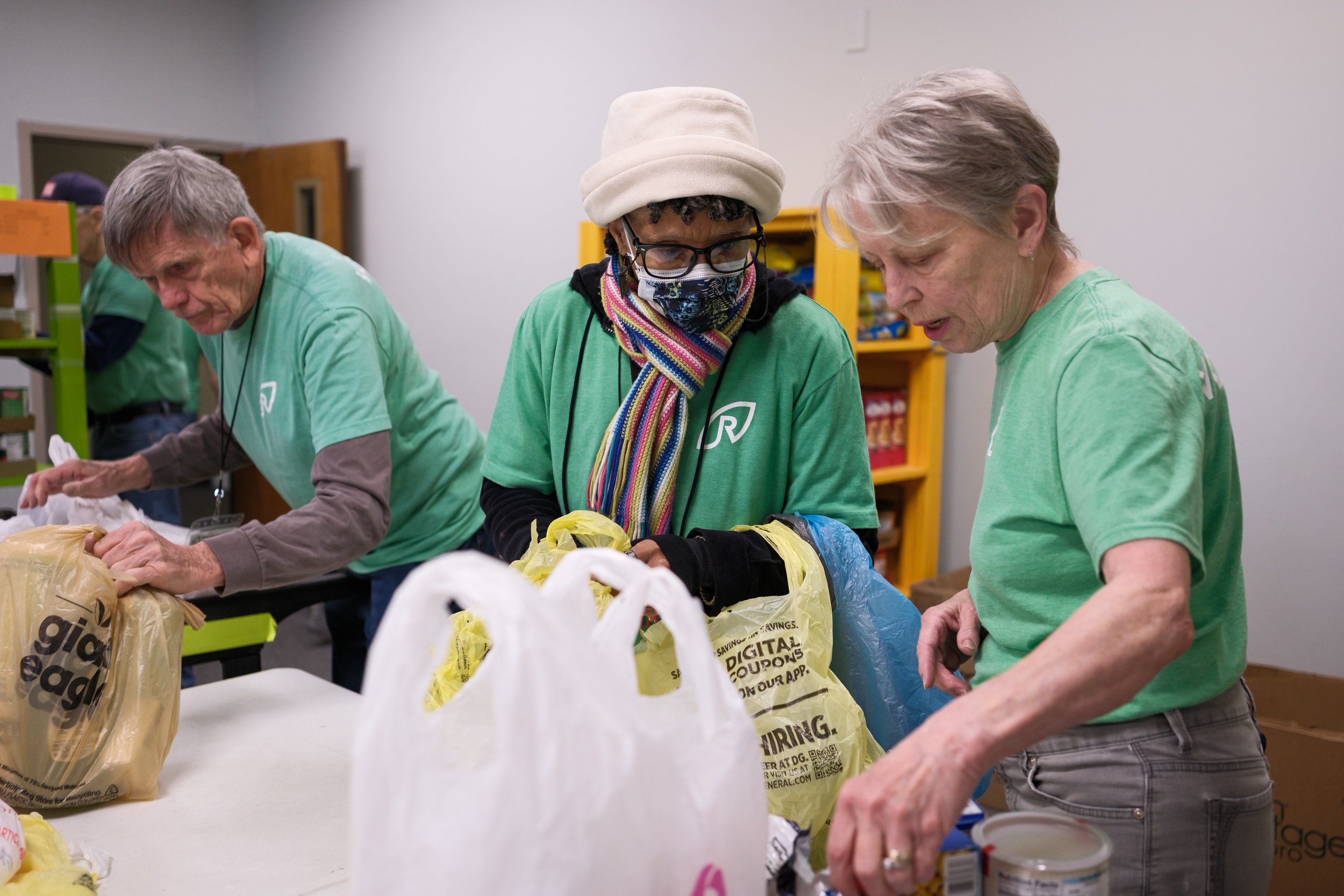 Three women, two elderly and one middle-aged, packaging food around a table, with plastic bags and canned goods in front of them, indoors in a community space.