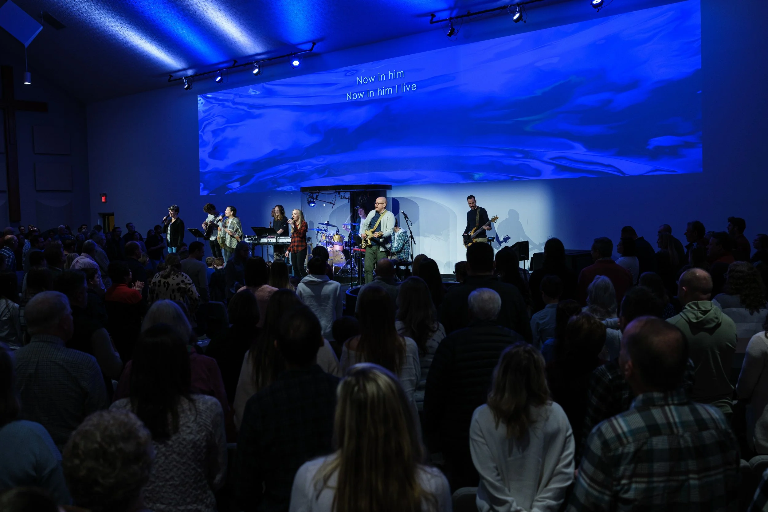 A large indoor gathering with a stage where a band is performing. The band includes multiple singers, guitar players, and a drummer. The audience is facing the stage, and the background features a large screen with blue visuals and the lyrics "Now in him I live" projected on it.