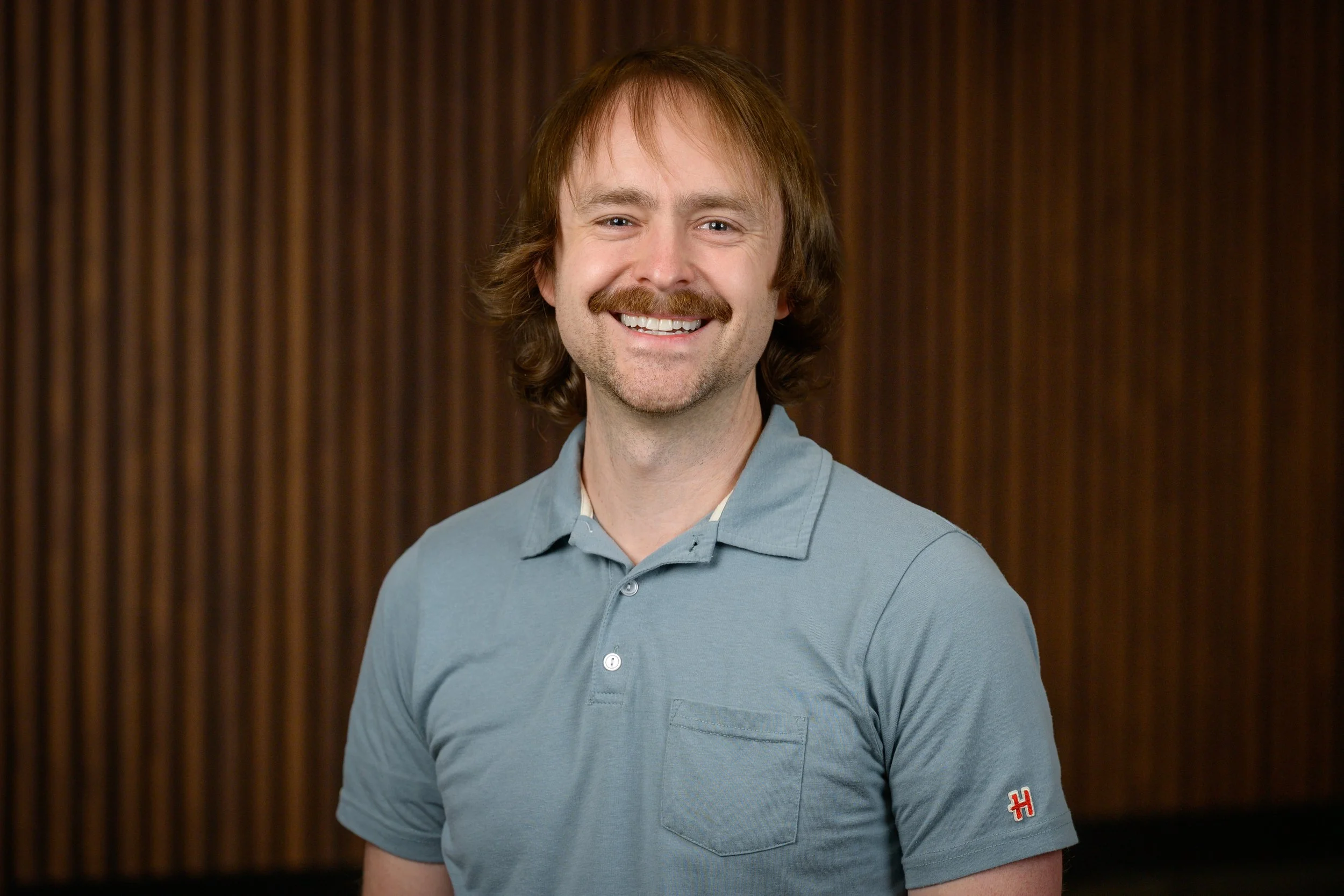 A young man with red hair, a mustache, and light skin, smiling and wearing a light blue polo shirt, standing in front of a dark wooden panel background.
