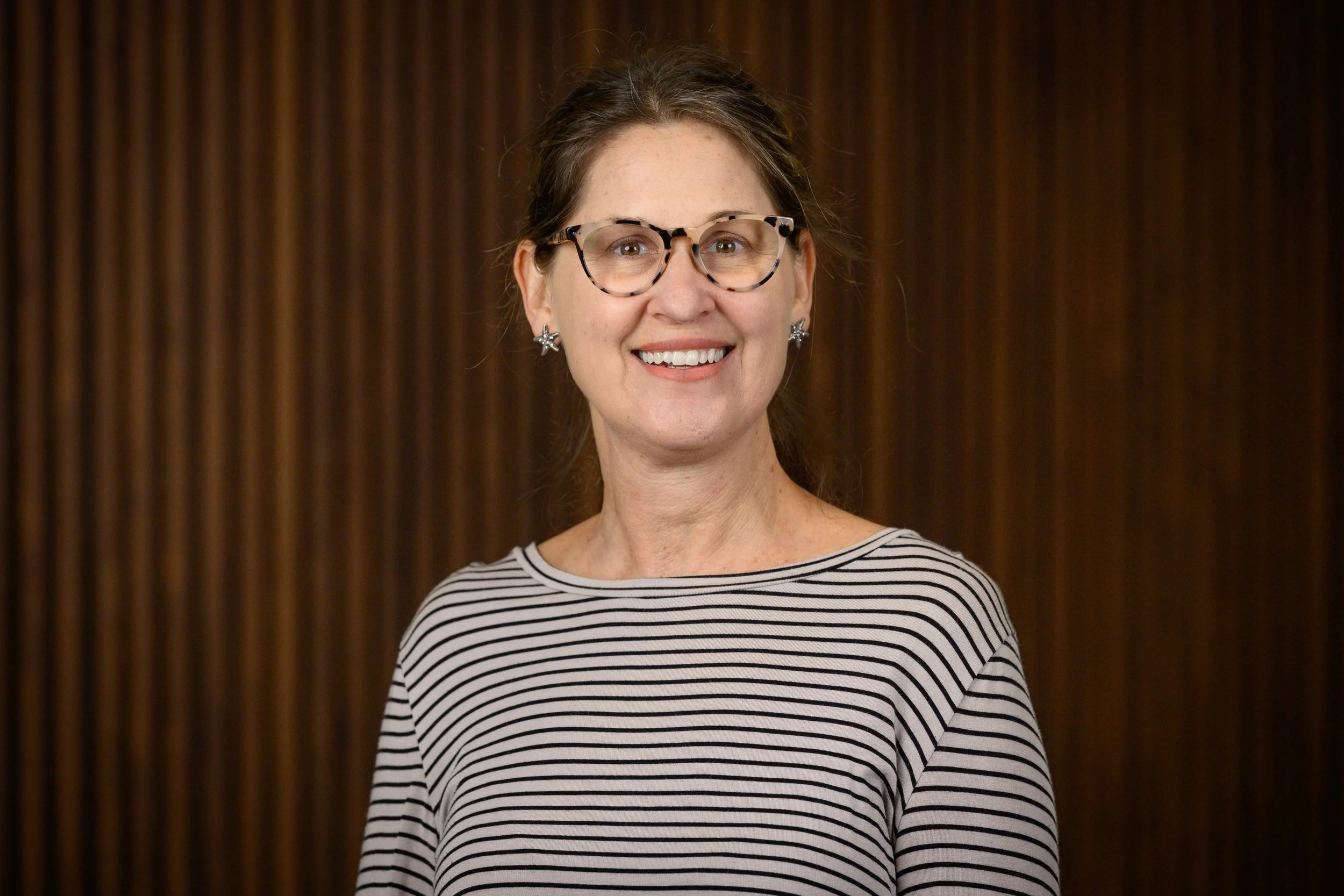 A woman smiling, wearing glasses and a black-and-white striped shirt, standing in front of a wooden background.