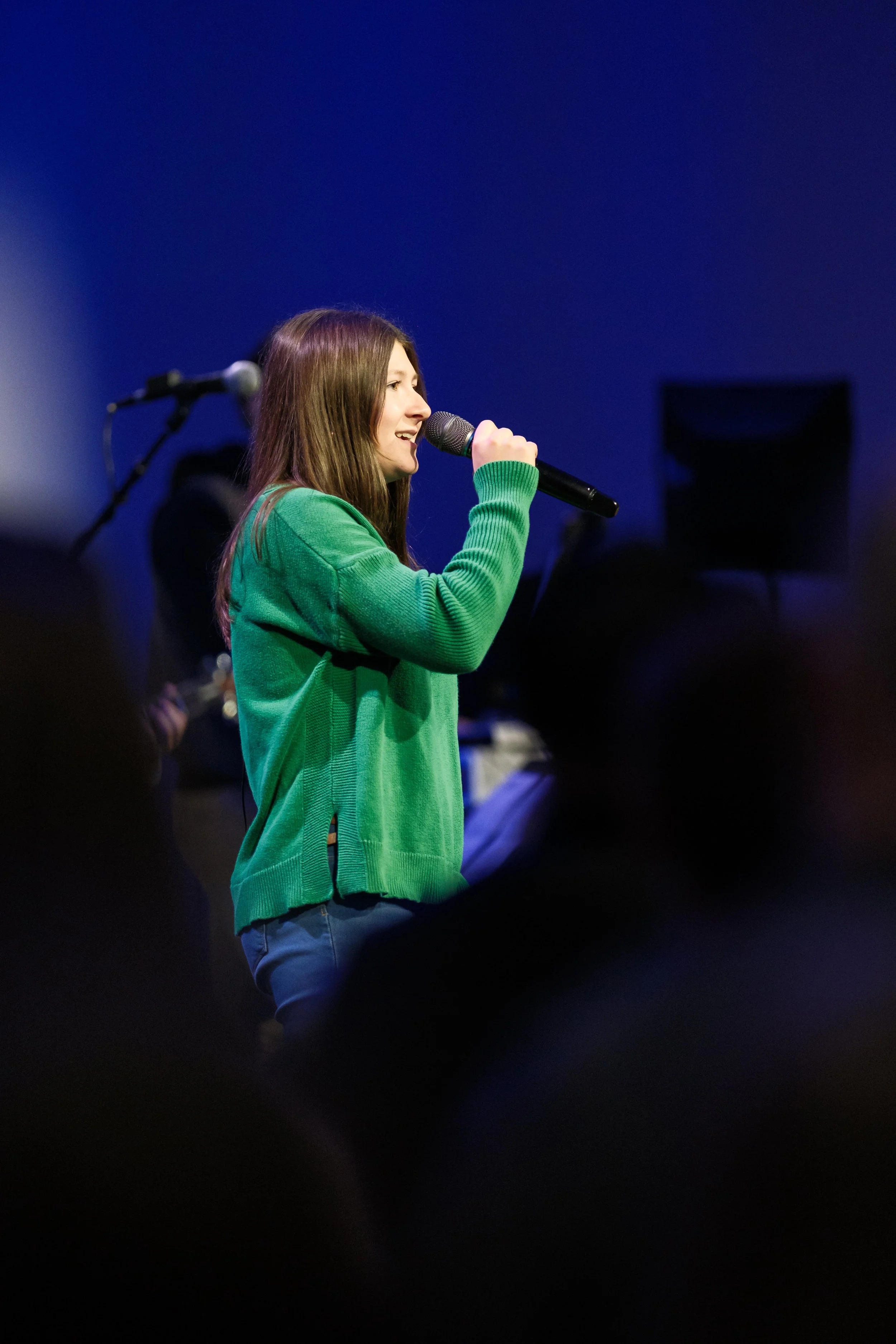 A young woman with long brown hair wearing a green sweater, singing into a microphone on stage with a dark blue background.