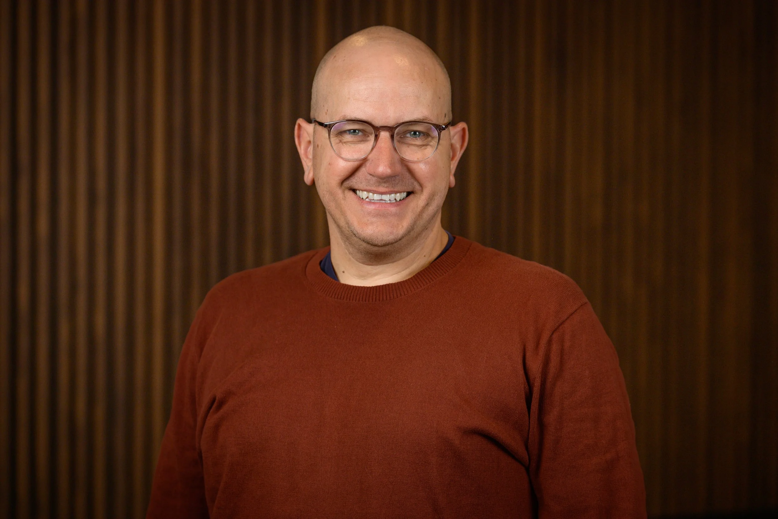 A smiling bald man with glasses wearing a red sweater, standing in front of a wooden wall.