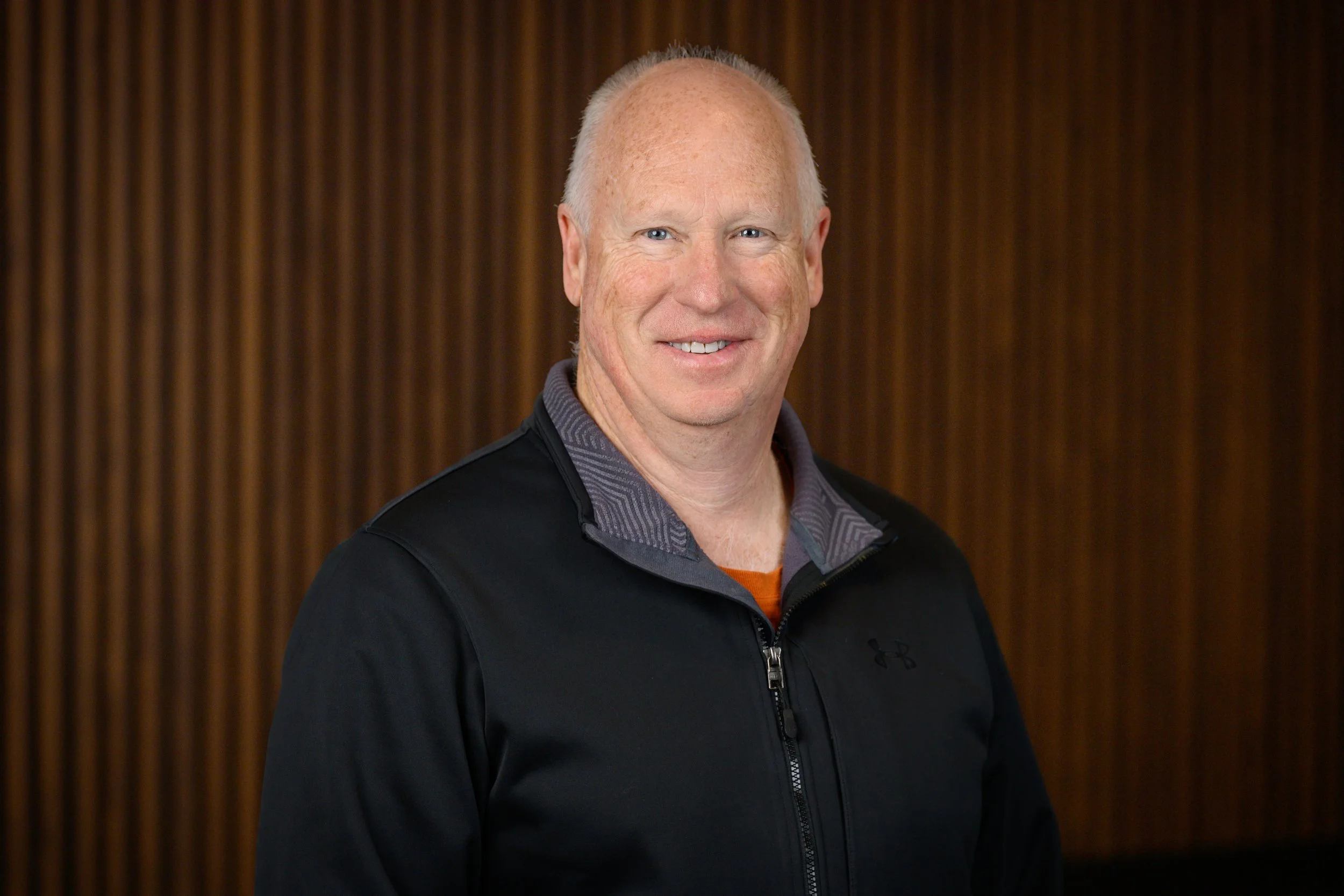 A smiling middle-aged man with short light-colored hair, wearing a black jacket over a gray collared shirt, standing in front of a wooden panel background.