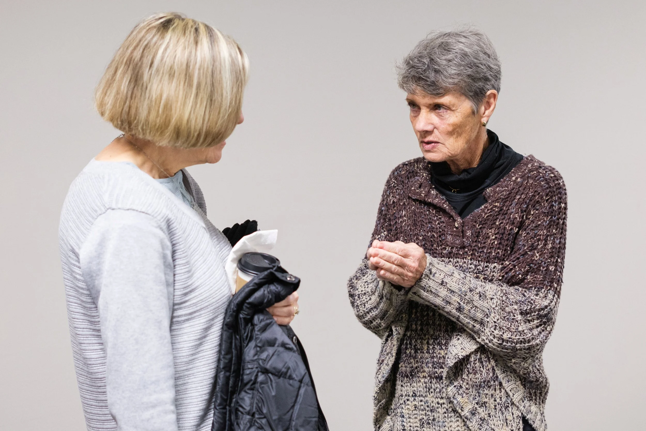 Two women engaged in conversation, one holding a coffee cup, indoors with a plain wall background.