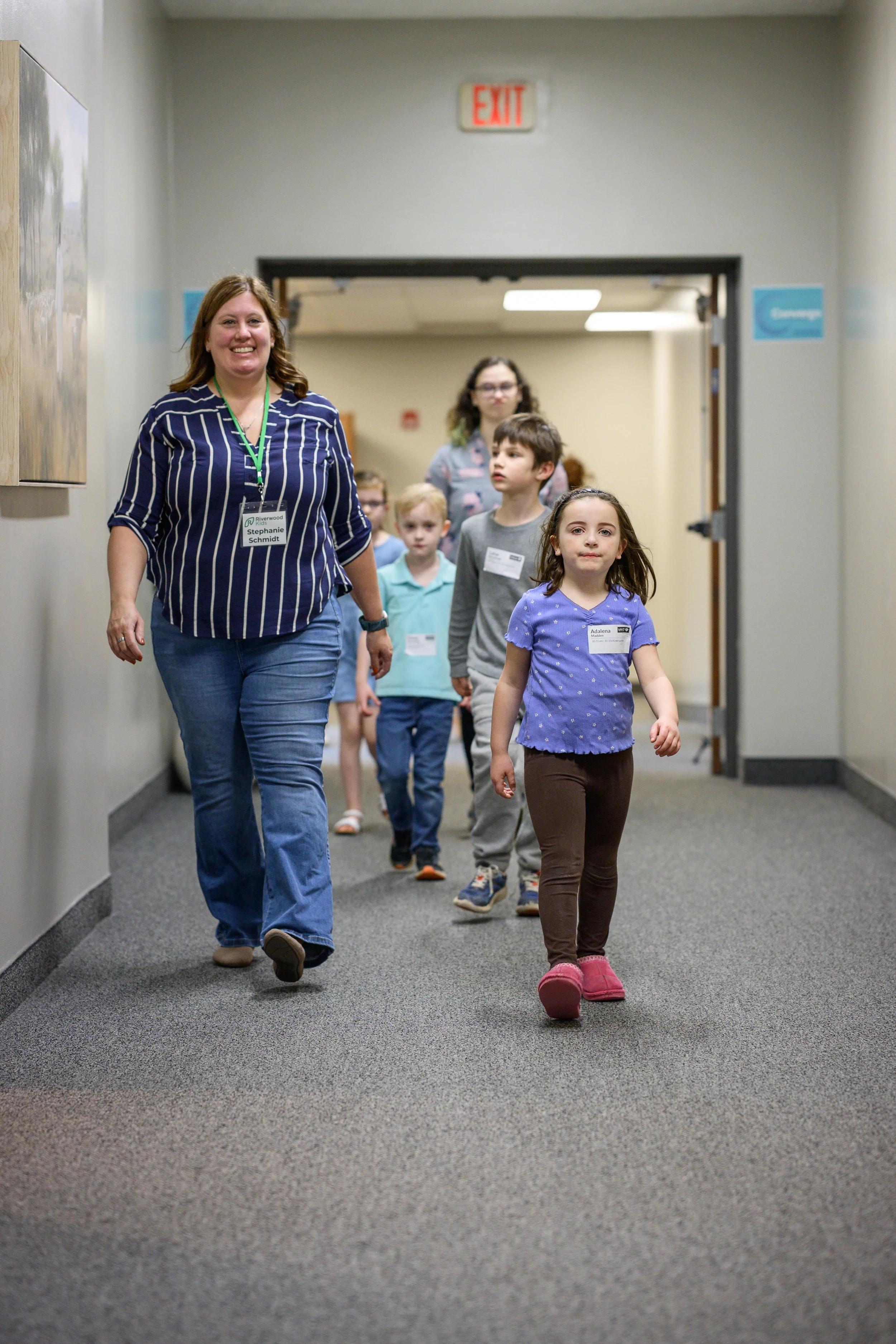 A woman leading a group of children down a hallway, all wearing name tags and walking confidently, with a bright exit sign above the doorway.