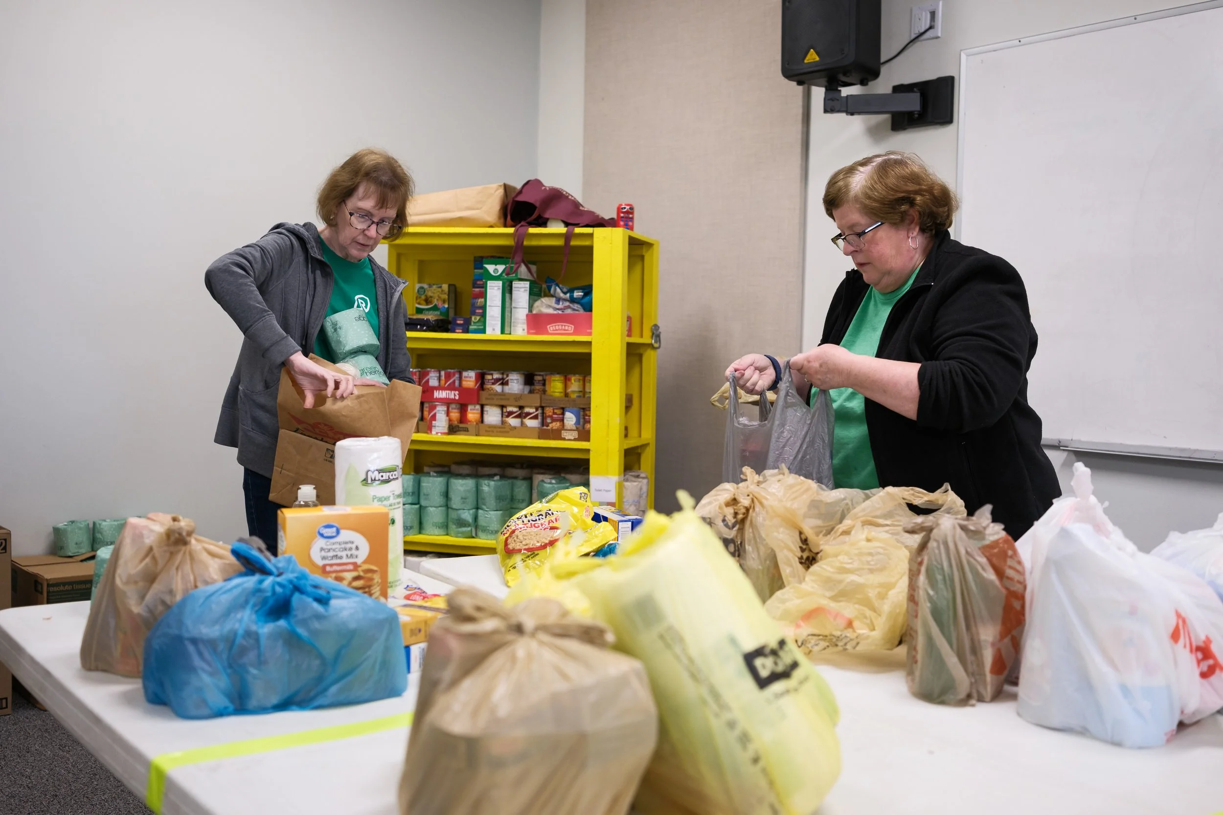 Two women packing food items at a table, with shelves of canned goods in the background.