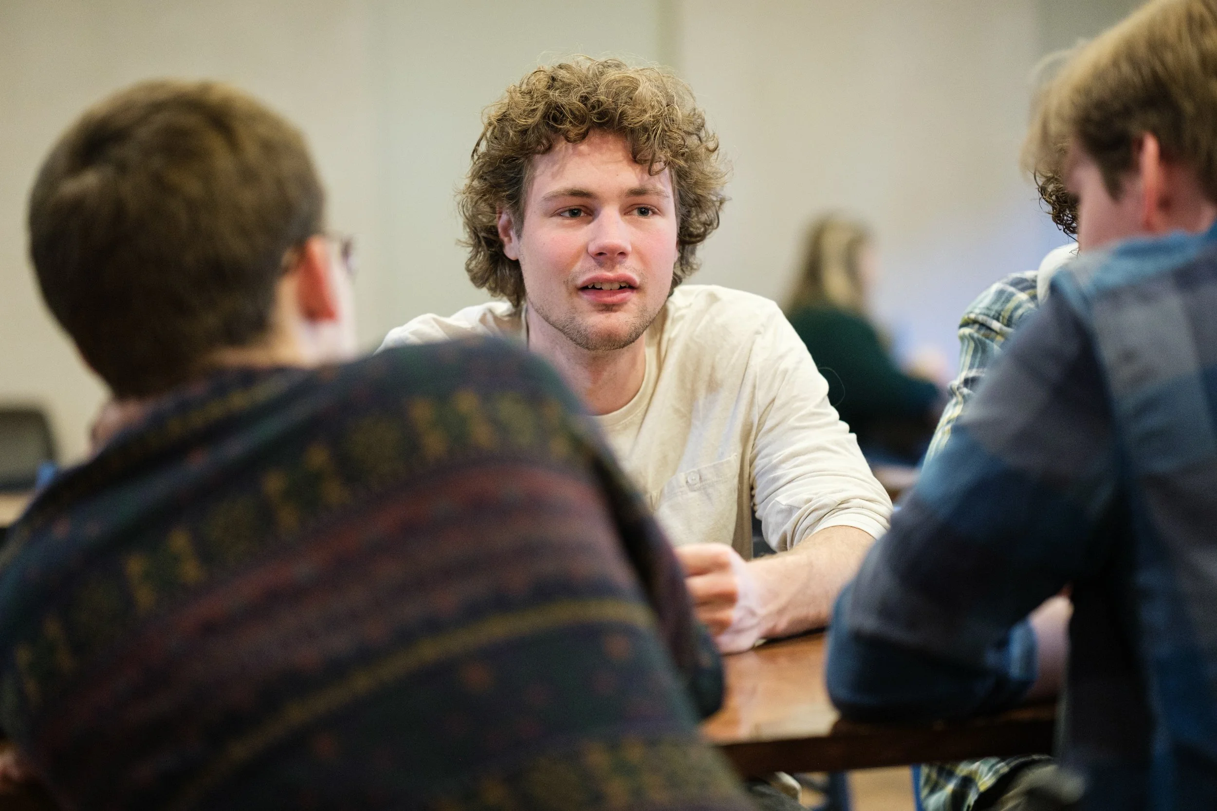 A young man with curly hair engaging in conversation with two people across a table in a casual indoor setting.