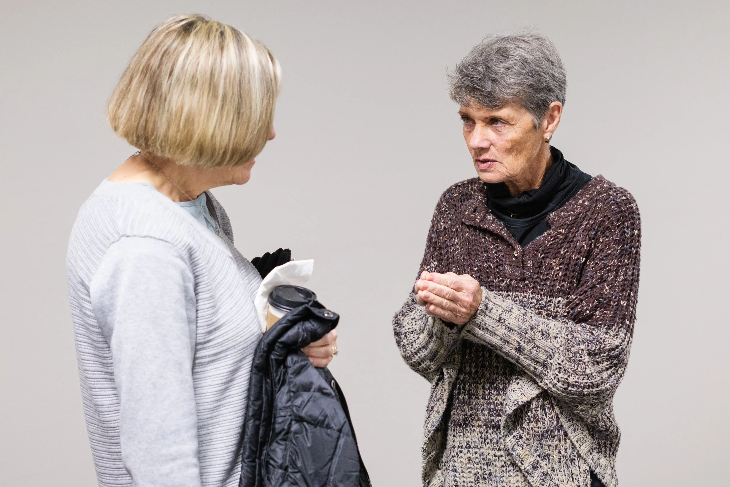 Two elderly women engaged in a conversation, one with blonde hair holding a coffee cup and the other with gray hair clasping her hands, wearing sweaters, standing against a plain background.