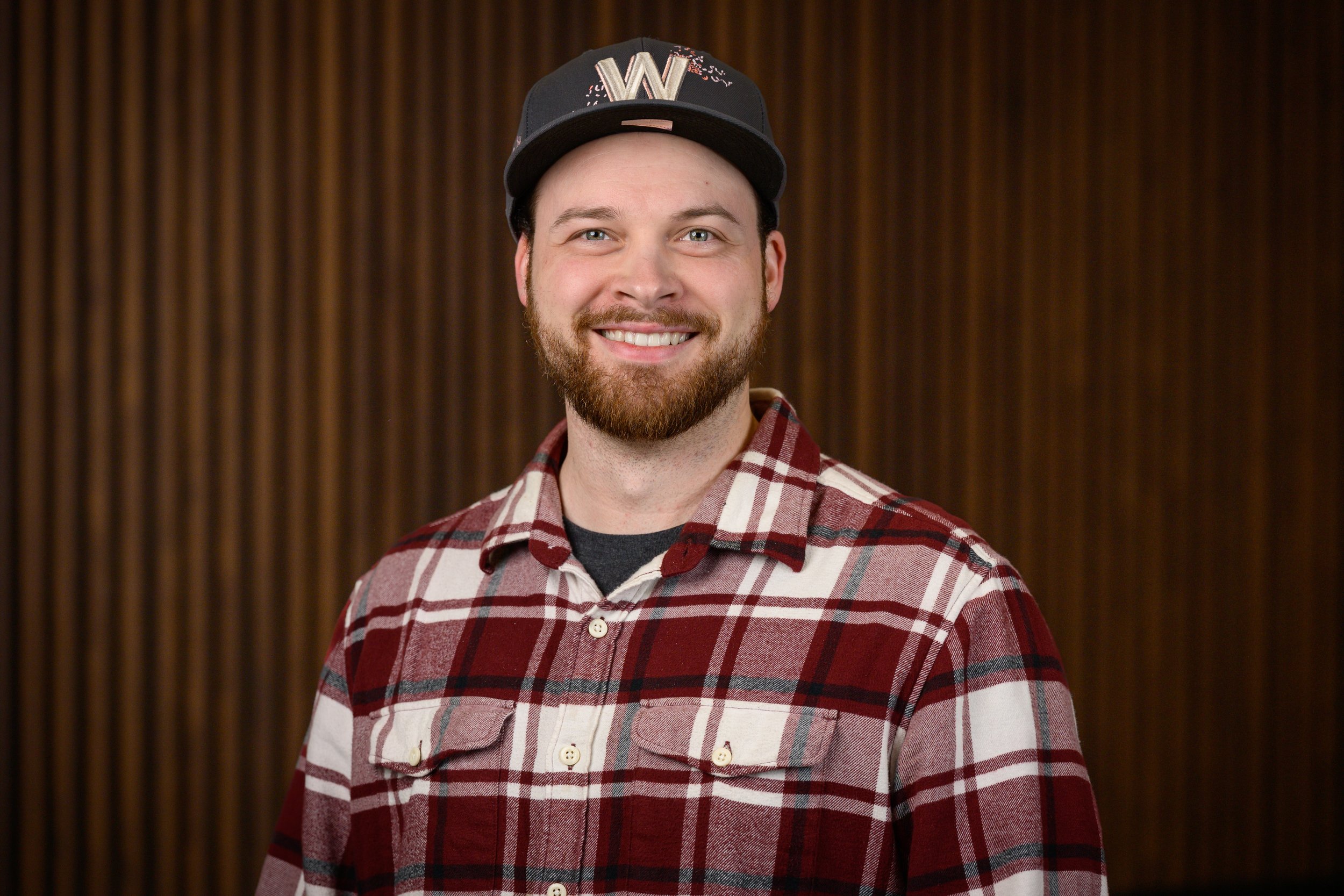 A smiling man with a beard wearing a red plaid shirt and a black baseball cap with a 'W' on it, standing in front of a brown striped background.