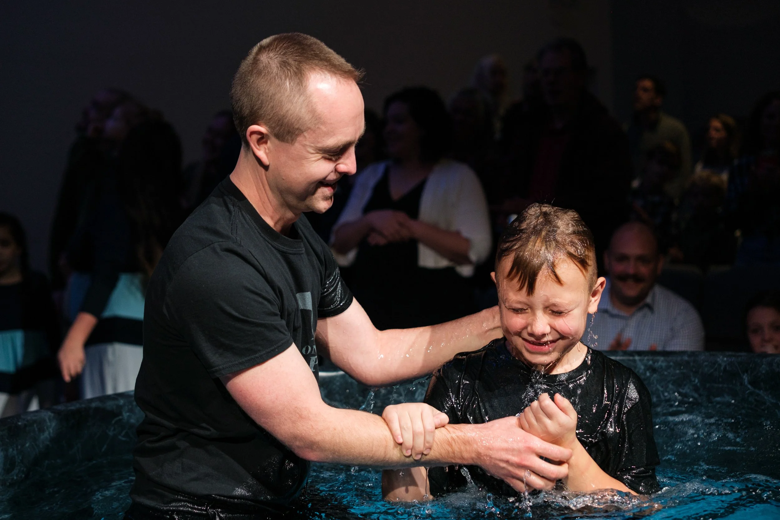 Man helping a young boy during a baptism ceremony in a church. The boy is crying and the man is holding his hands. There are people watching in the background.