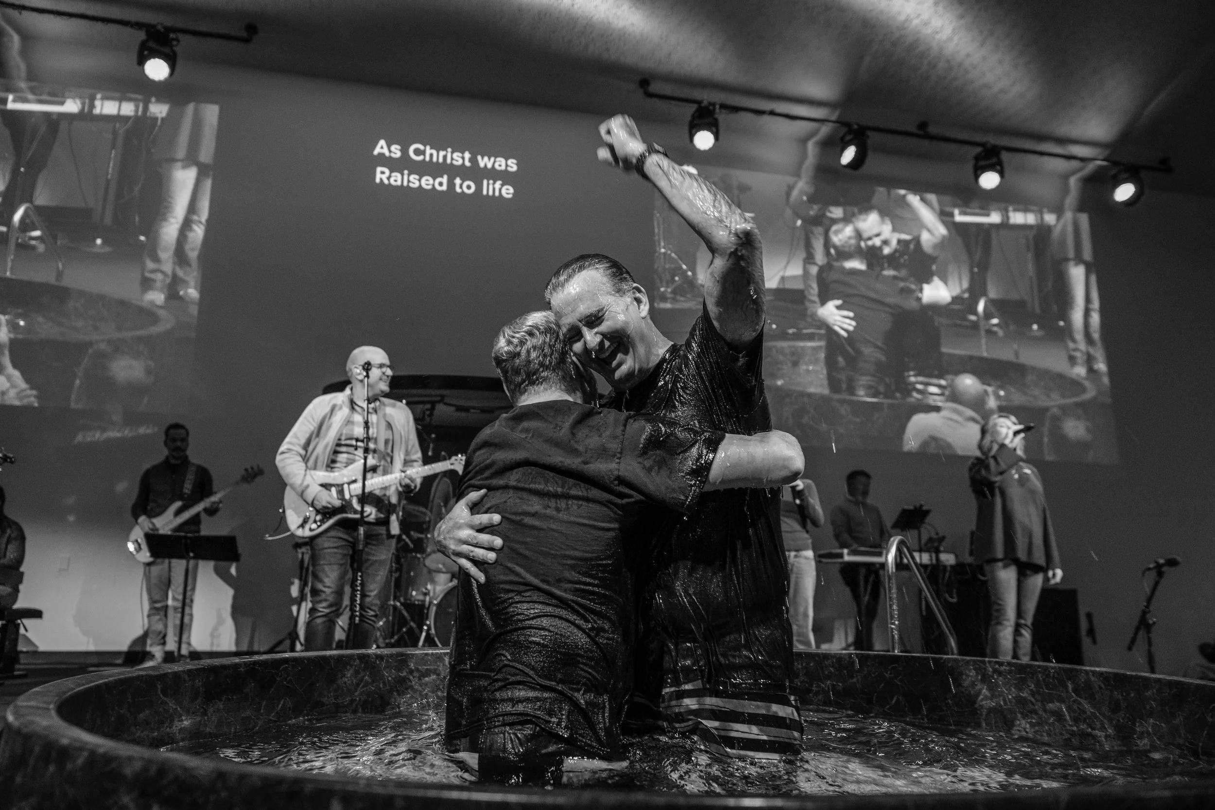 A black and white image of two people embracing in a baptismal pool during a baptism ceremony, with a church band playing in the background and a large screen displaying the words, "As Christ was Raised to life."