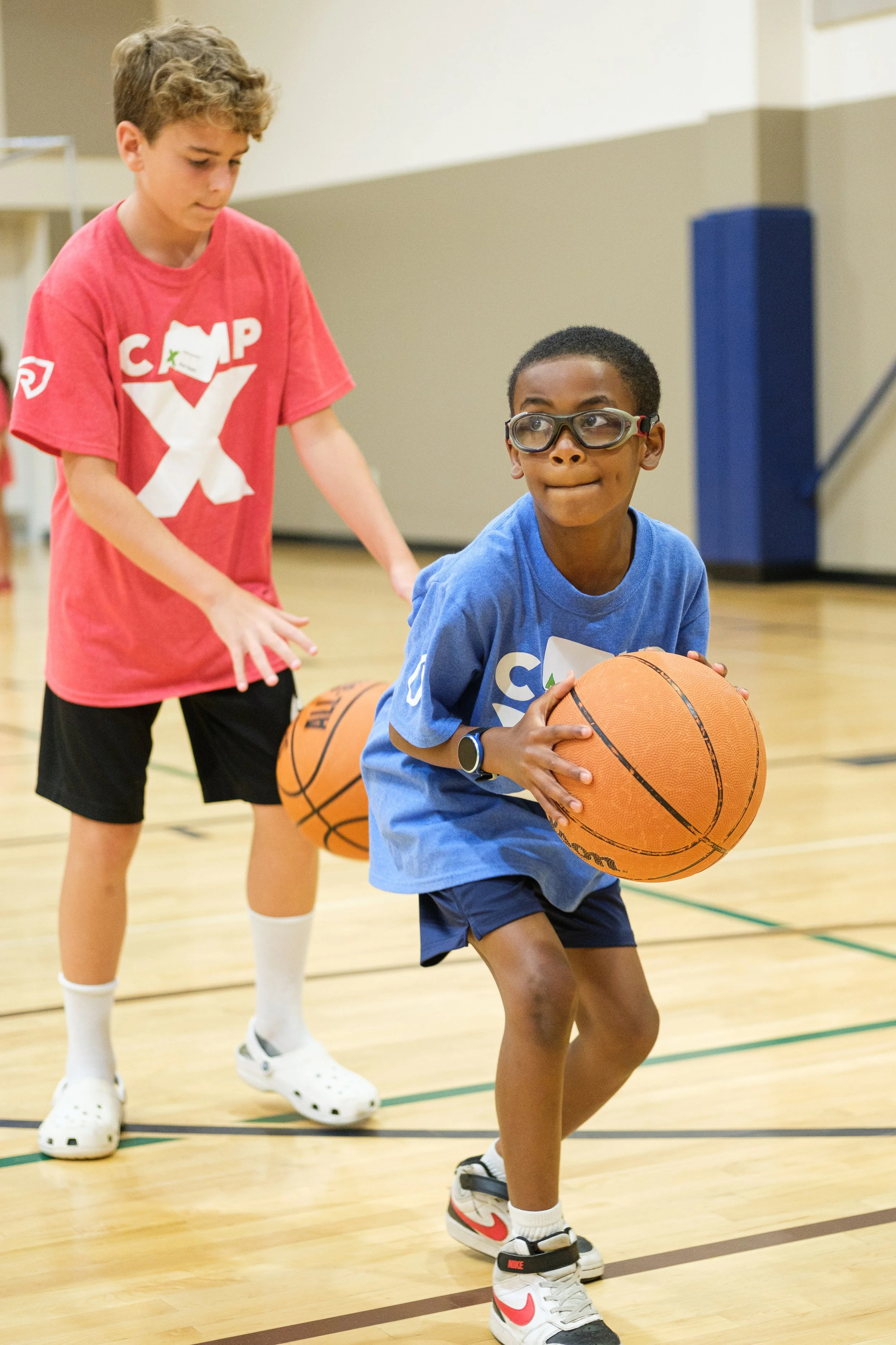 Two boys playing basketball in an indoor gym. The boy in the foreground is wearing glasses, a blue t-shirt, and Nike sneakers, holding a basketball. The boy in the background is wearing a red t-shirt and white Crocs, standing behind him.
