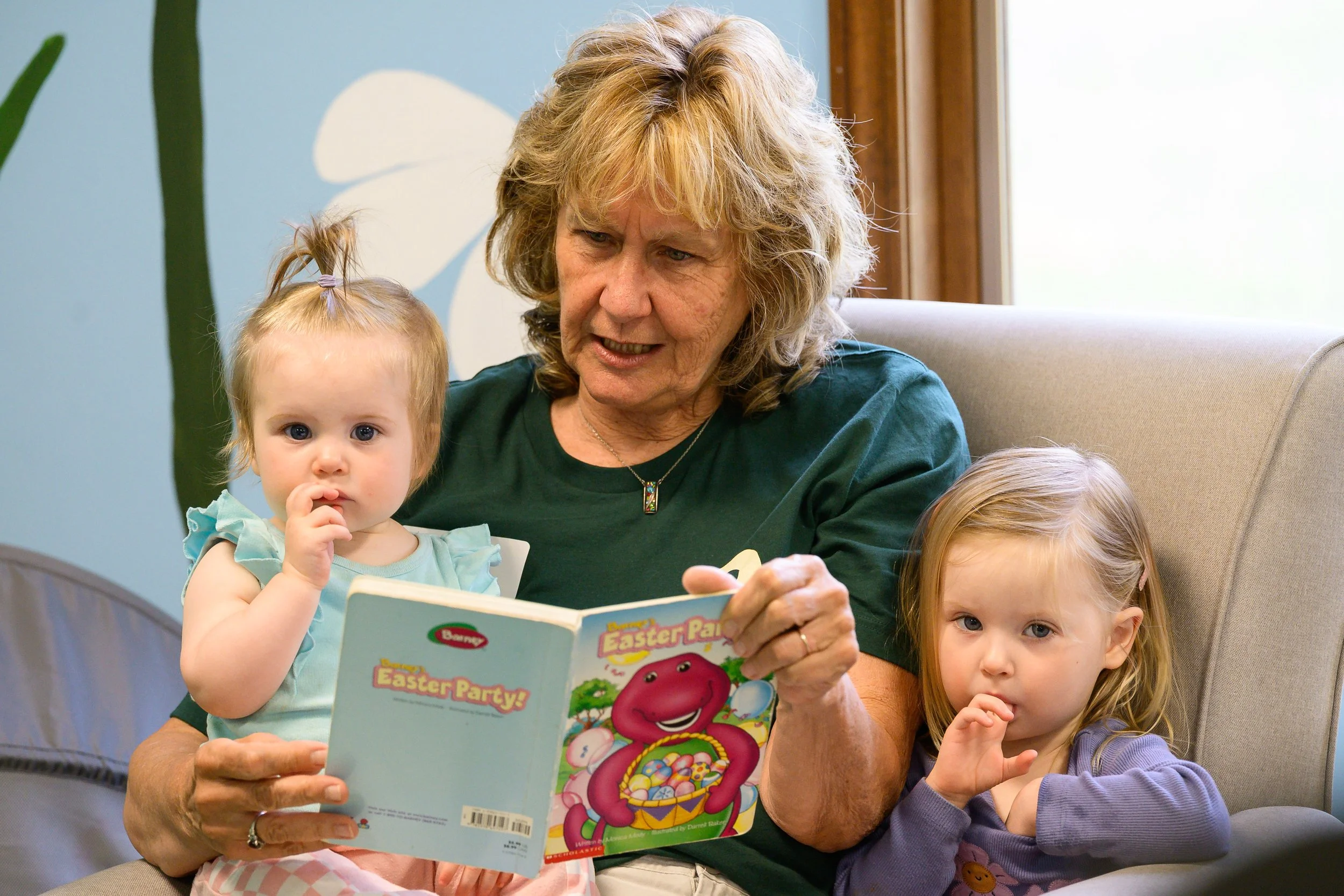 An older woman with curly blonde hair reading a children's book titled "Easter Party!" to two young girls with brown hair, one of whom has pigtails, sitting on her lap in a room with light blue walls and a window.