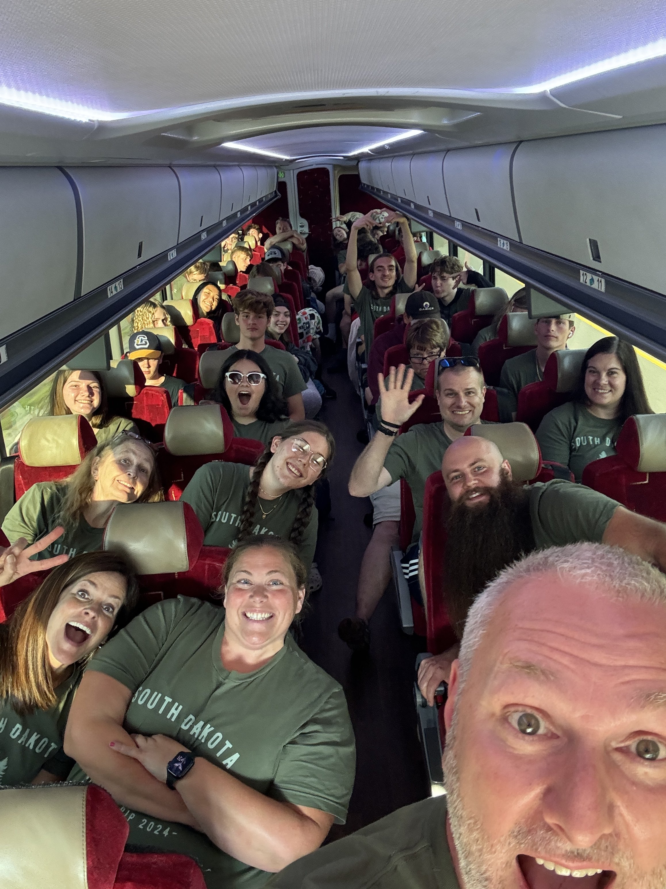 Group of smiling people on a bus, some wearing green shirts with 'South Dakota' written on them, taking a selfie together.