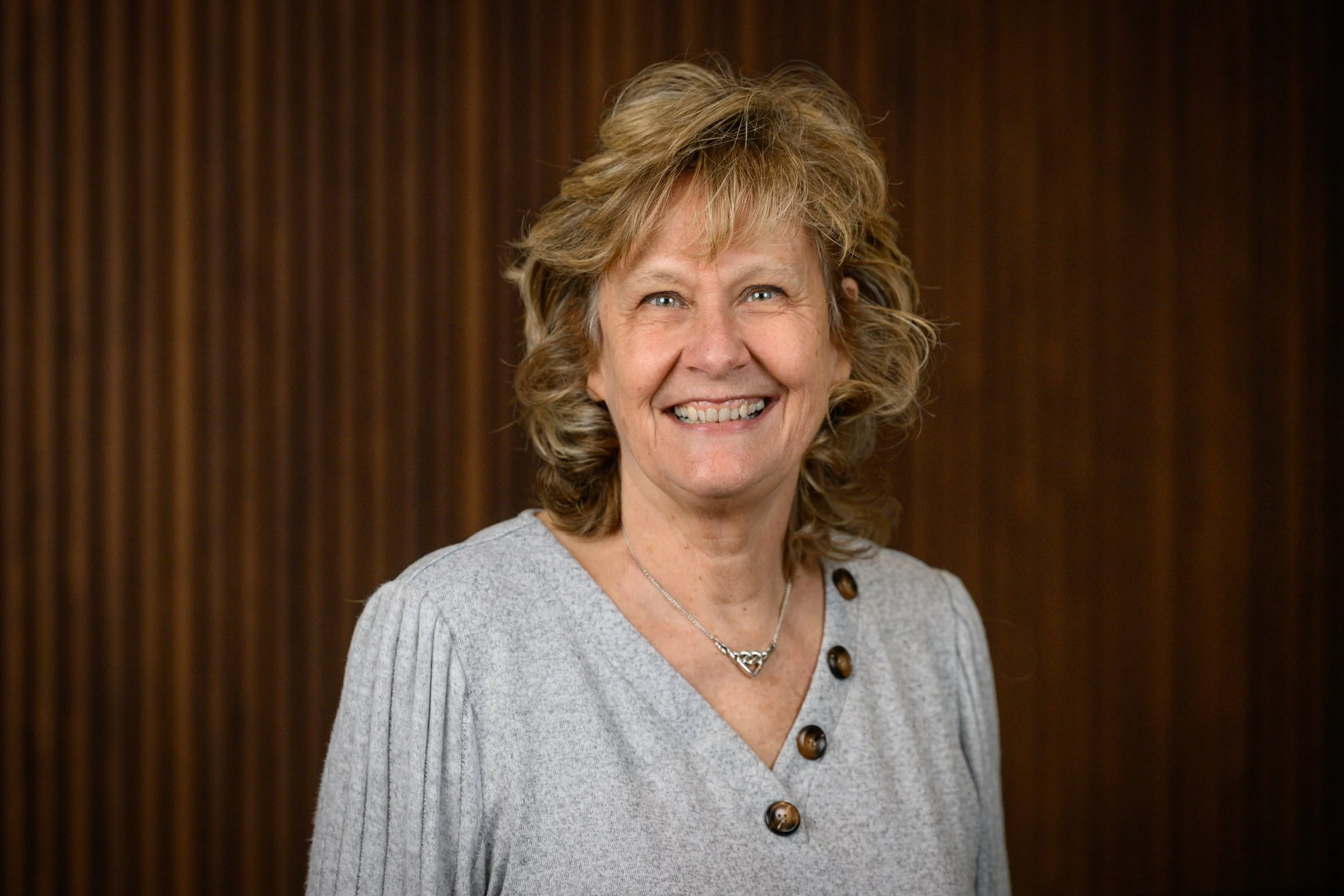 A smiling woman with shoulder-length curly blonde hair, wearing a gray sweater with large buttons and a silver necklace, standing against a wood-paneled background.