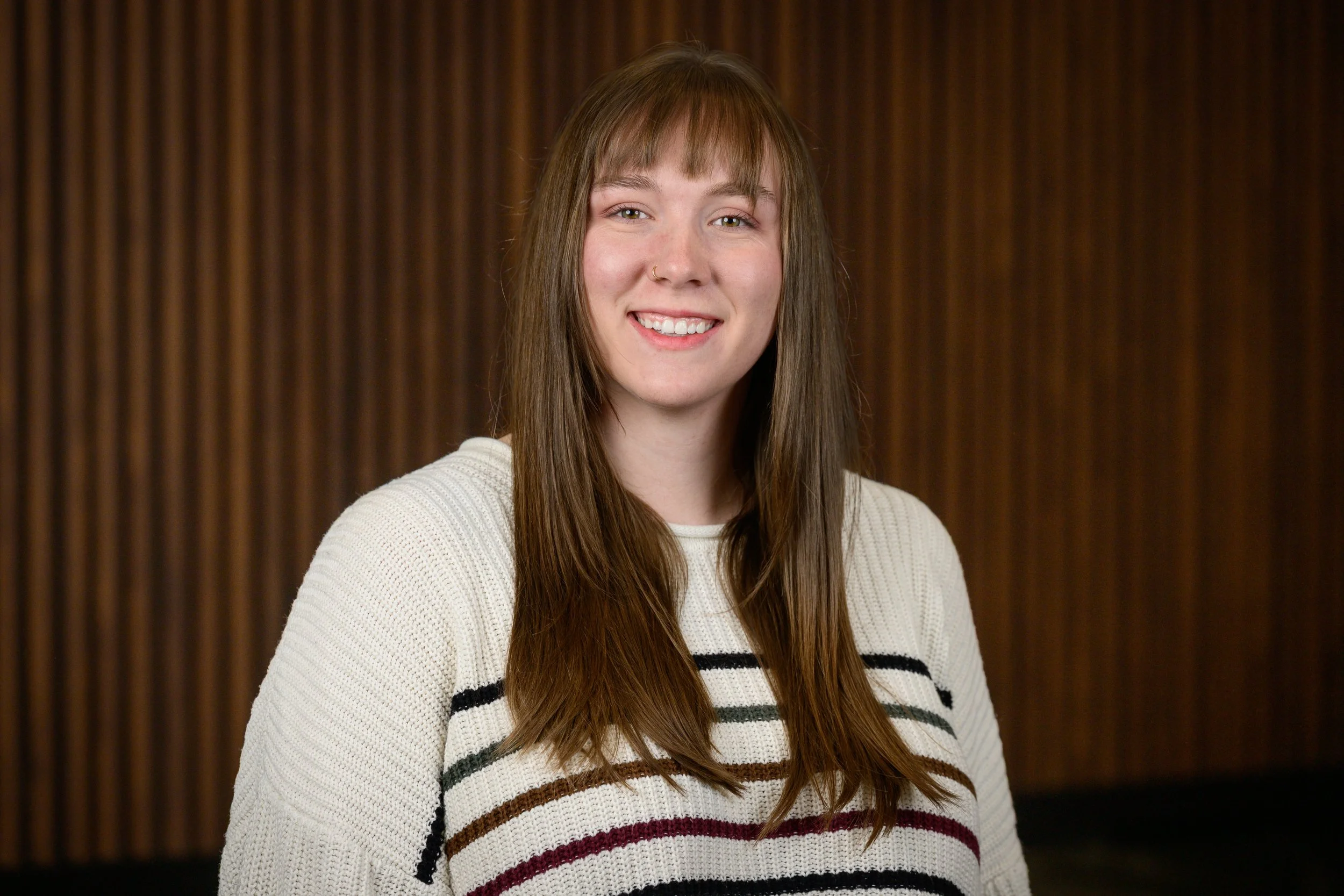 Young woman with long brown hair, wearing a cream and striped sweater, smiling in front of a wooden background.