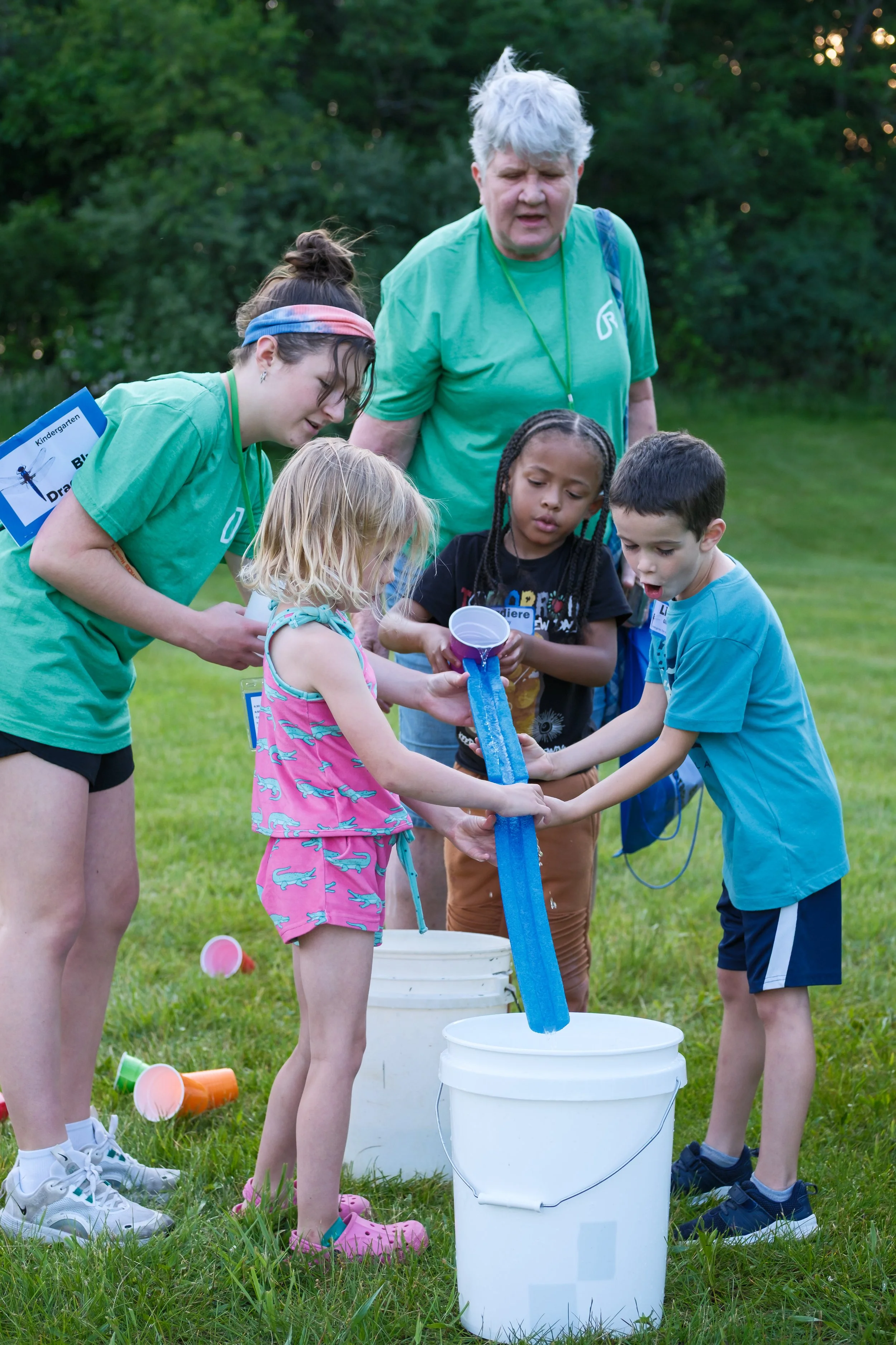 Group of children and an older woman participating in a water activity outdoors on a grassy field, with lush green trees in the background. The children are holding and pouring water through a long blue tube into a white bucket, while the woman watches. The children are dressed in casual clothes and the woman is in a green t-shirt.