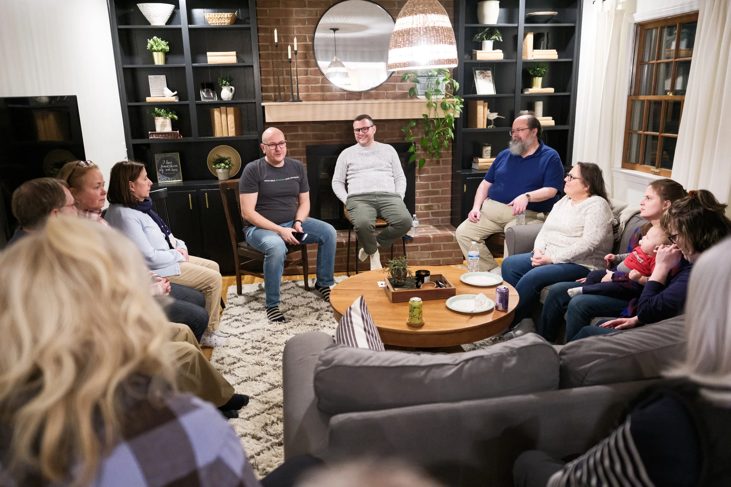 People sitting on couches and chairs in a living room, engaged in a group discussion or meeting, with a brick wall, decorative shelves, and a round mirror in the background.