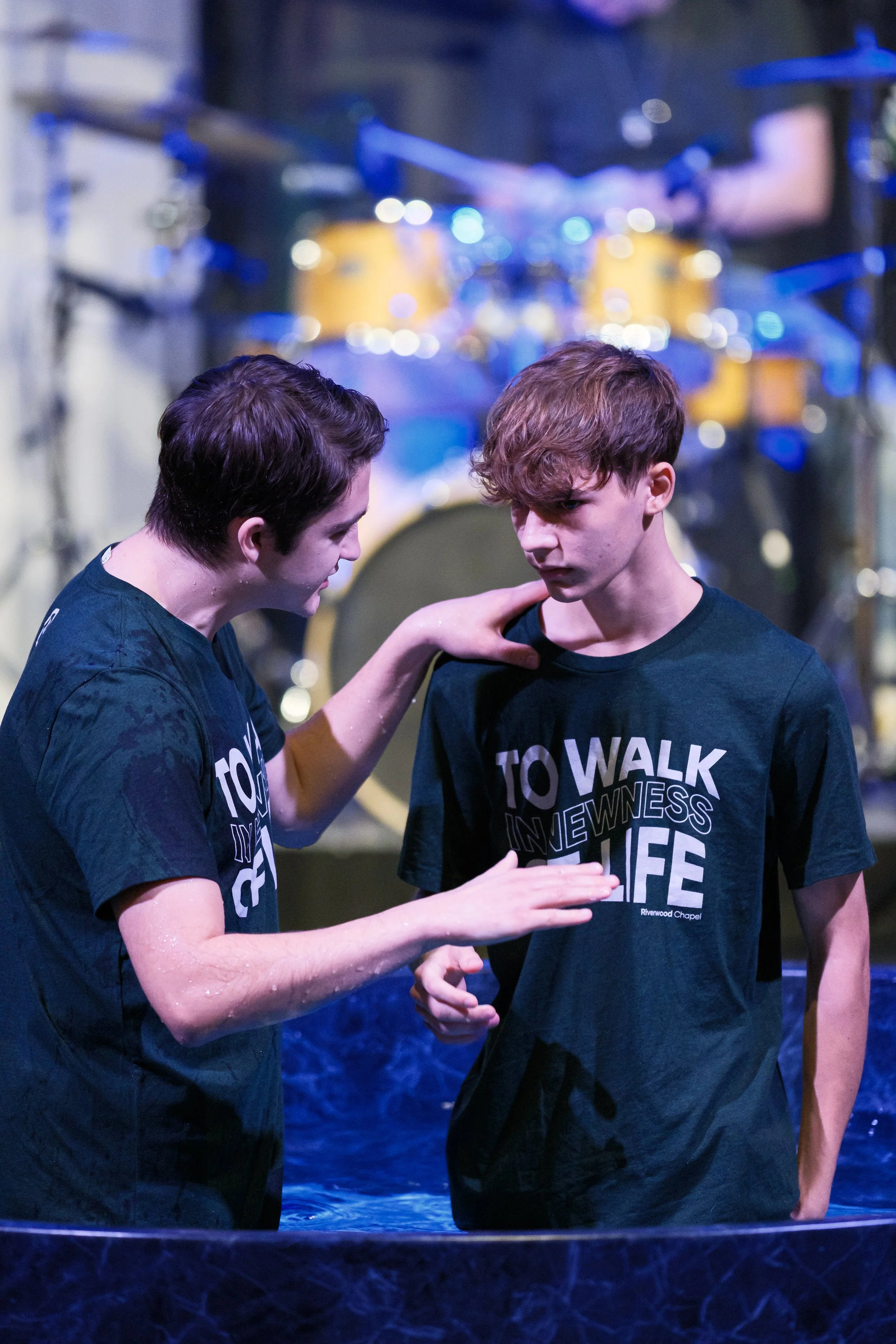 Two young men in black T-shirts with the words "TO WALK IN WITNESS IN LIFE" are standing in a shallow water baptism pool, with one of them placing his hand on the other's shoulder while the other appears to be getting baptized. The background shows stage lighting and musical instruments.