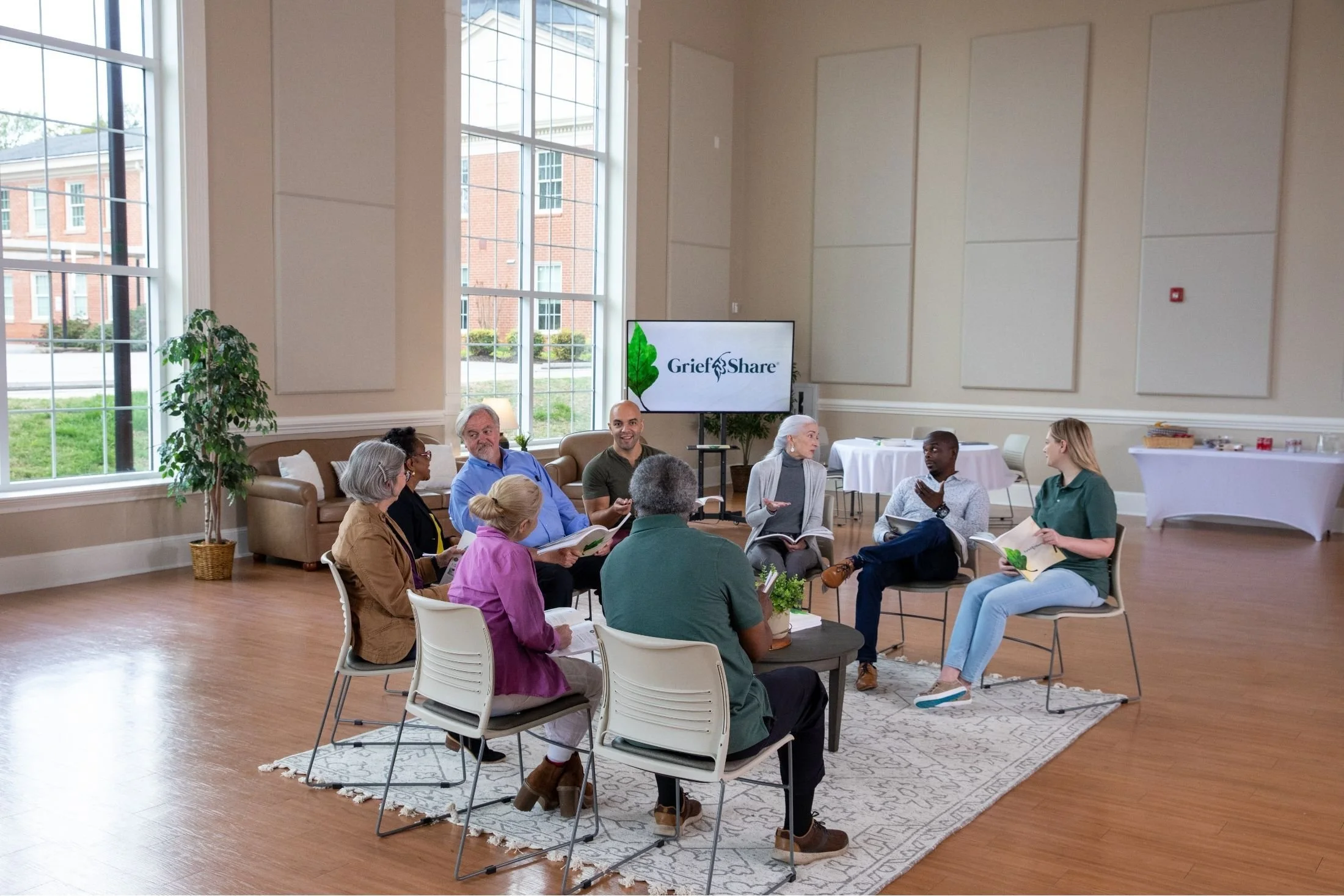 Group of people seated in a circle participating in a discussion or meeting in a bright room with large windows, a television displaying 'GriefShare', and some tables with refreshments in the background.