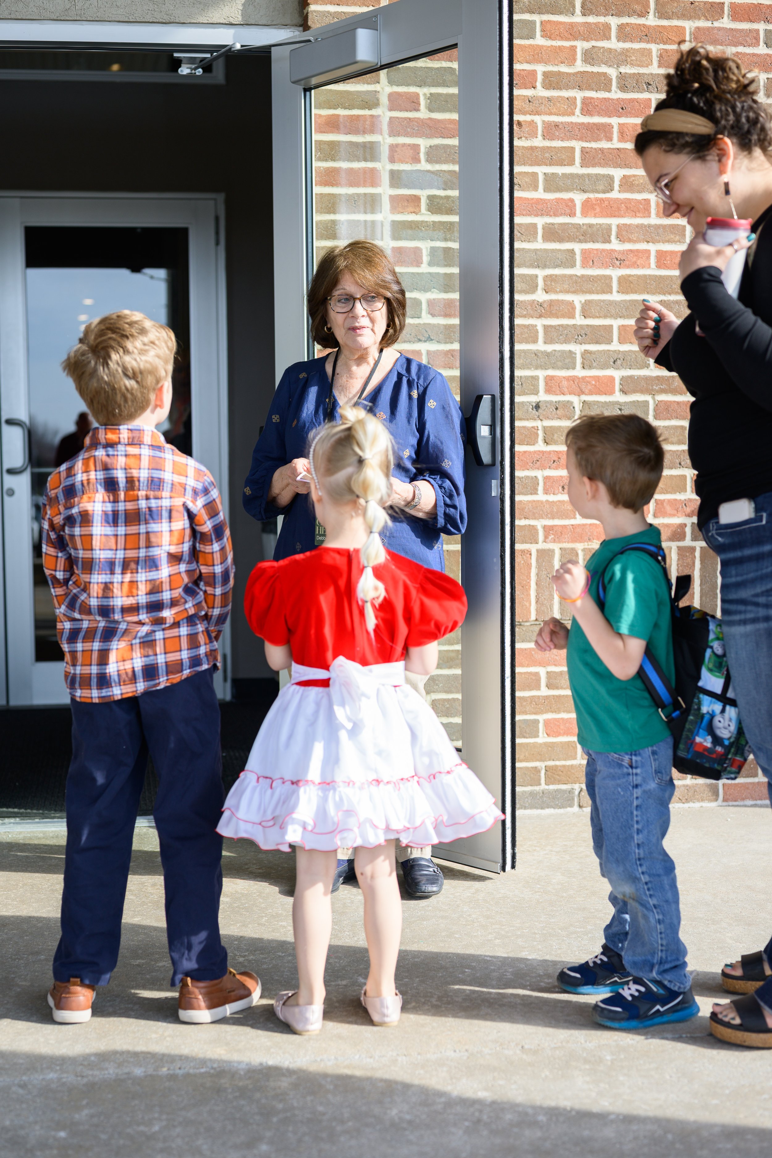 A woman standing outside a building, talking to four children who are queuing up. The children are dressed smartly, and one girl is in a red and white dress. The setting appears to be daytime near an entrance door.