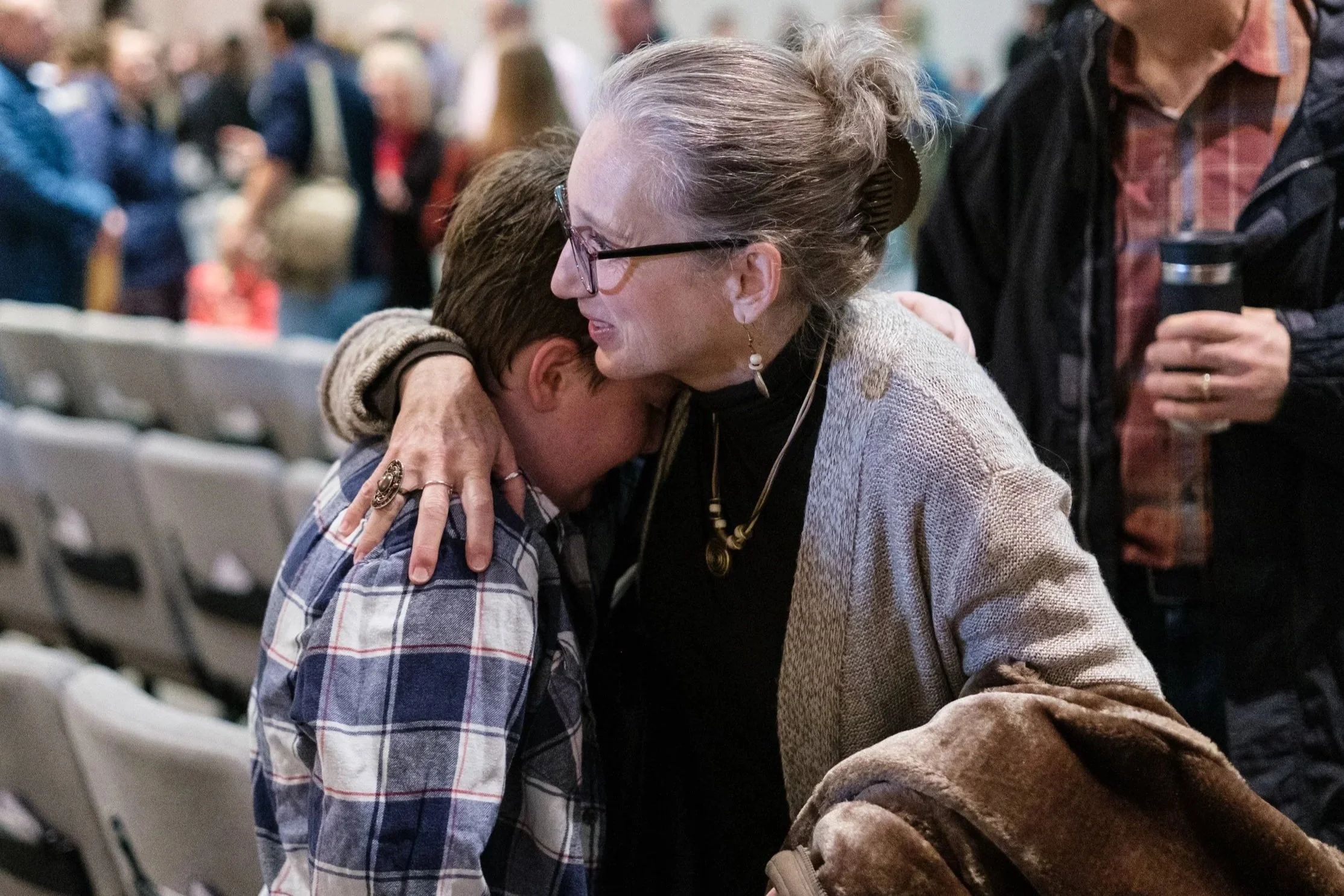 A woman with gray hair and glasses embraces a young boy in a checkered shirt, in a crowded indoor setting with blurred people in the background.