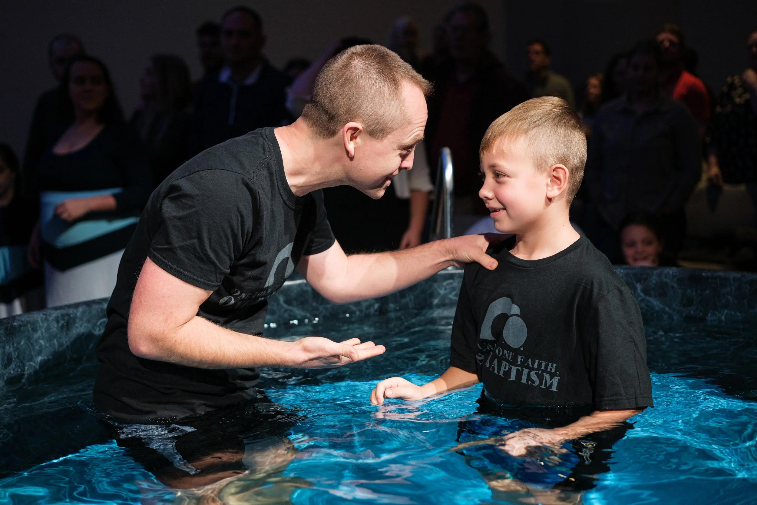 A man and a young boy standing in a baptismal pool, smiling and looking at each other during a baptism ceremony, with a group of people in the background.