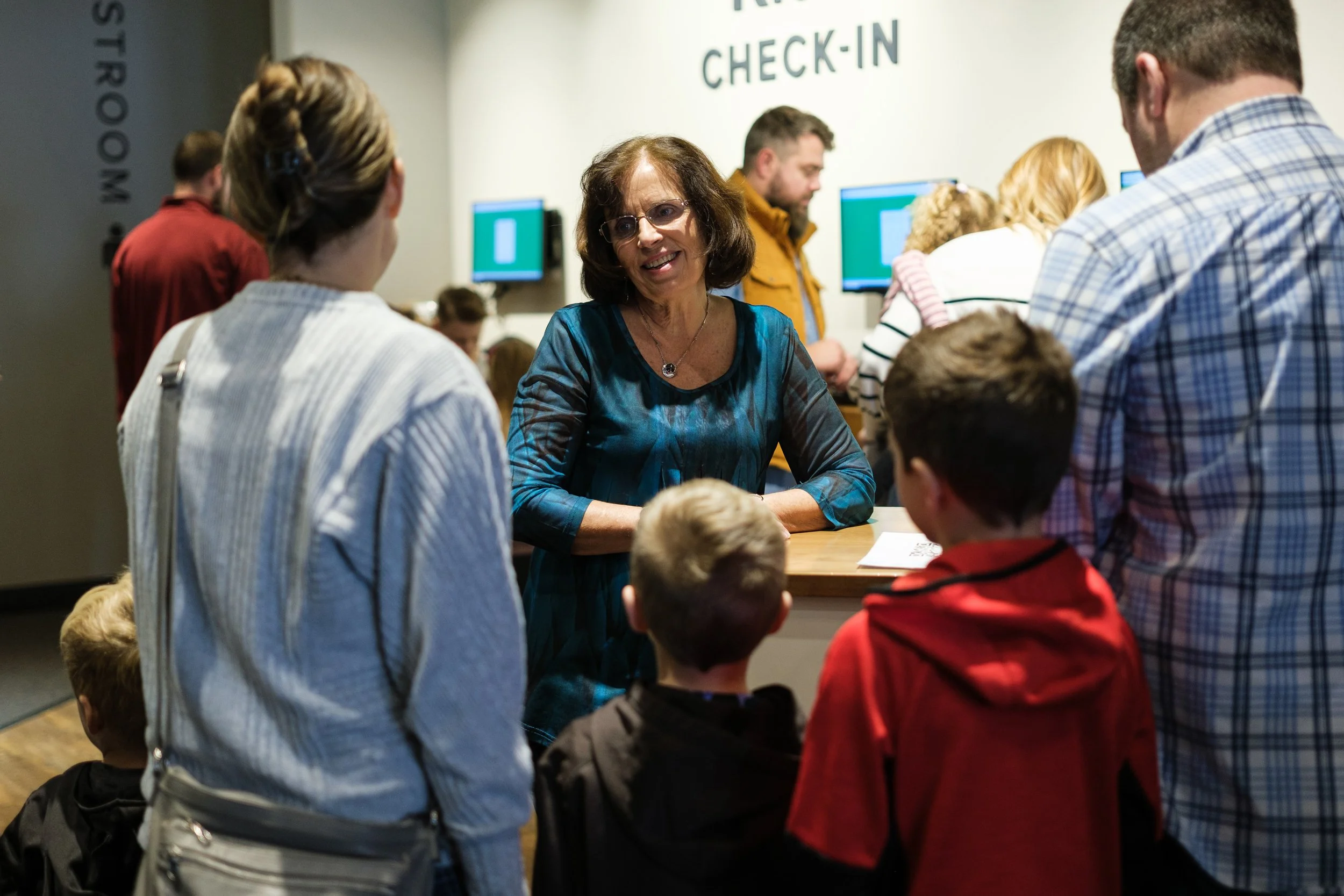 A group of people gathered around a woman behind a check-in counter at an airport. The woman is smiling and talking to the group. The background has monitors and a sign that reads 'CHECK-IN'.