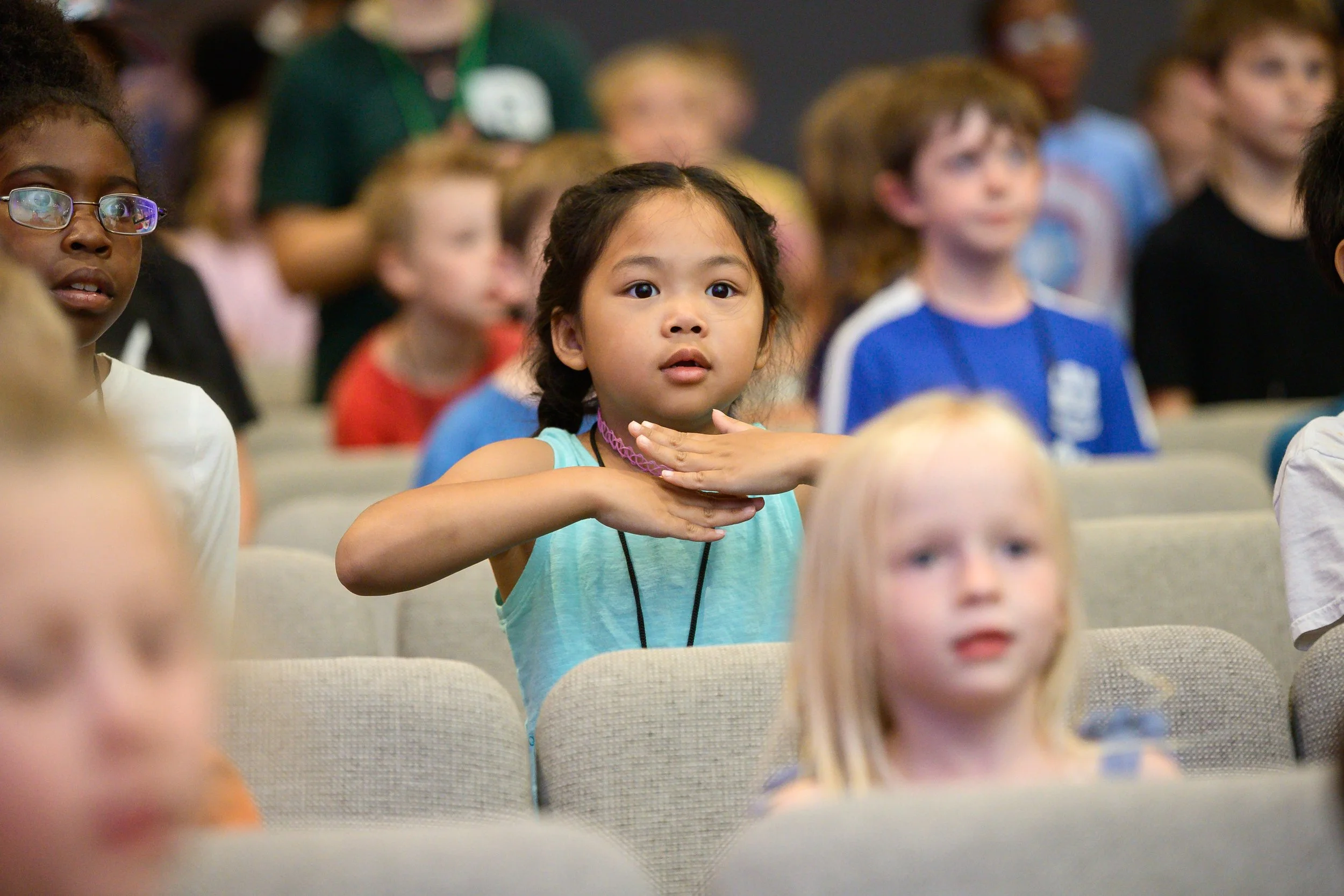 A young girl with dark hair in a braid, wearing a turquoise shirt and a purple choker, sitting in an auditorium among other children, some of whom are blurred in the background.