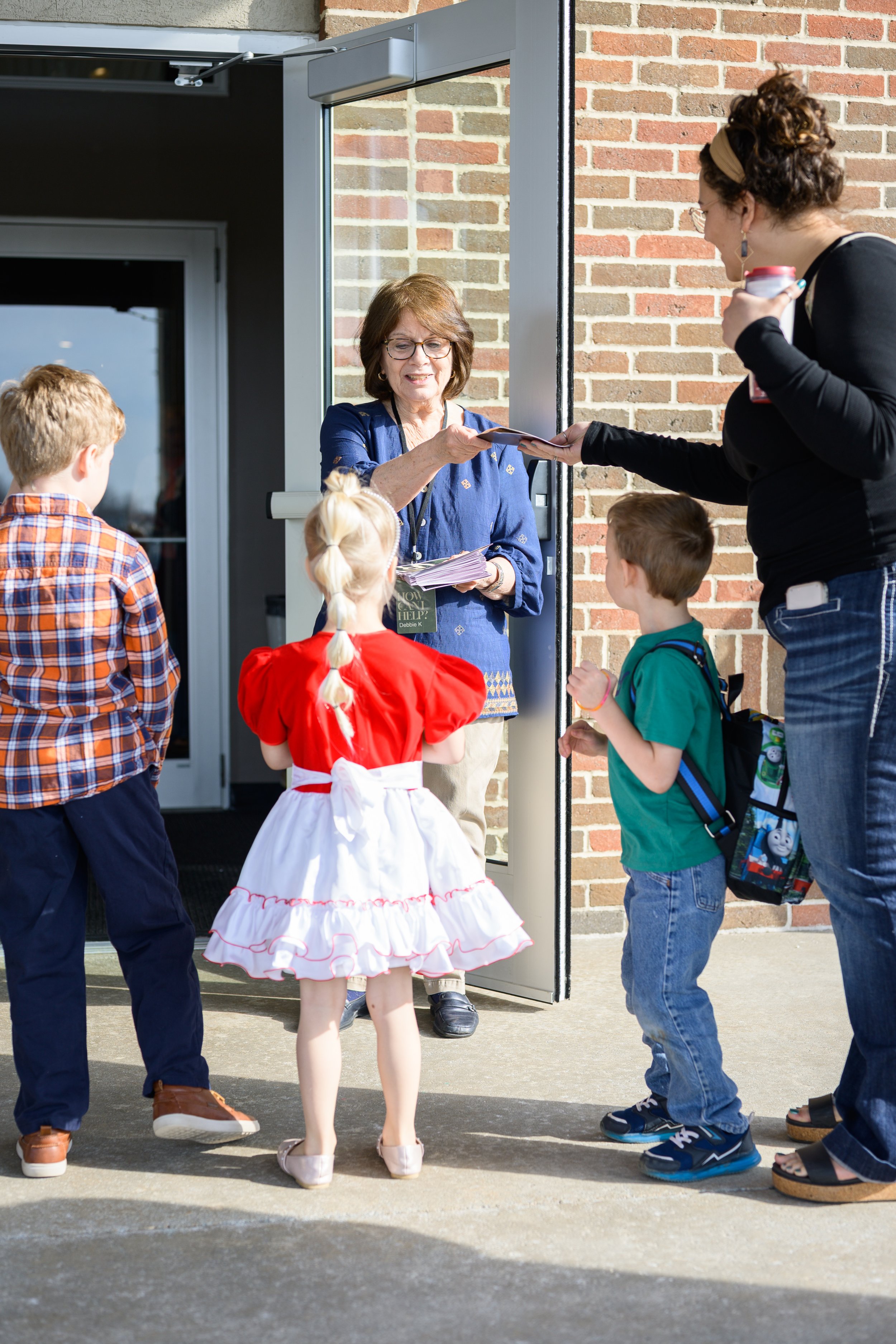 A group of children with backpacks and a woman in a black top handing out papers to a woman standing at the door of a building with a brick wall. It appears to be outside a school or community center.