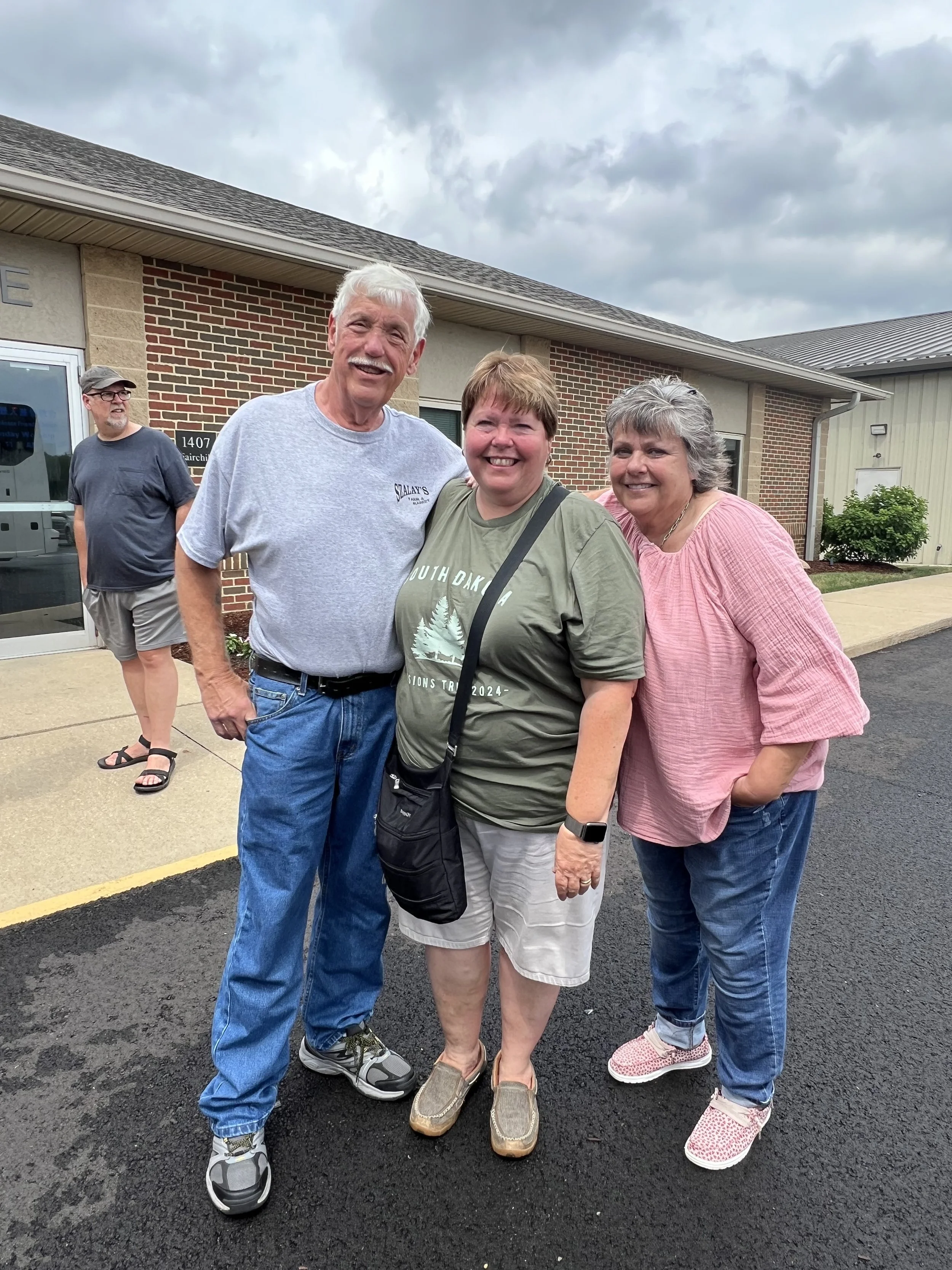 Group of four smiling adults standing outdoors in front of a brick building, with cloudy sky above.